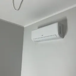 Biały klimatyzator marki AUX zamontowany na jasnej ścianie w rogu pomieszczenia, widoczny kabel zasilający przy suficie.