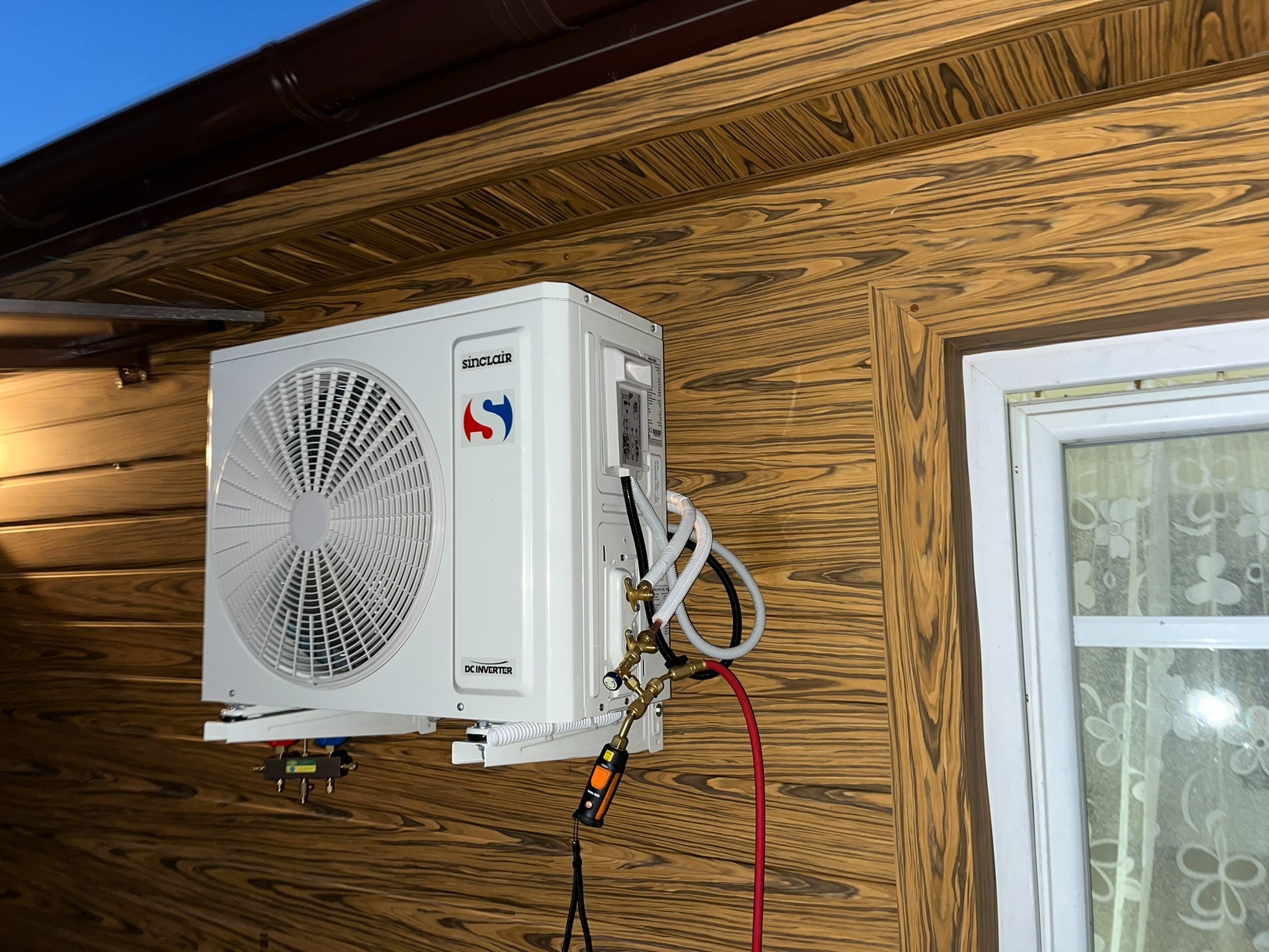 Zewnętrzna jednostka klimatyzacji Sinclair zamontowana na ścianie budynku z drewnianą okładziną, widoczne podłączenia rur i przewody, w tle fragment okna z firanką.