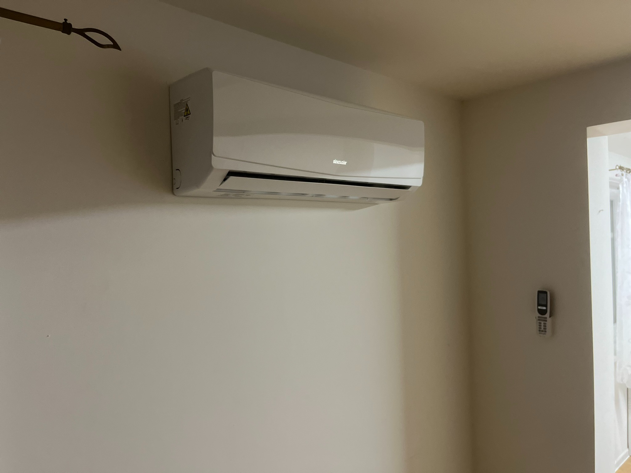 Biała klimatyzacja marki Sinclair zamontowana na ścianie w jasnym pomieszczeniu, widoczny pilot na ścianie obok i fragment karnisza w rogu.