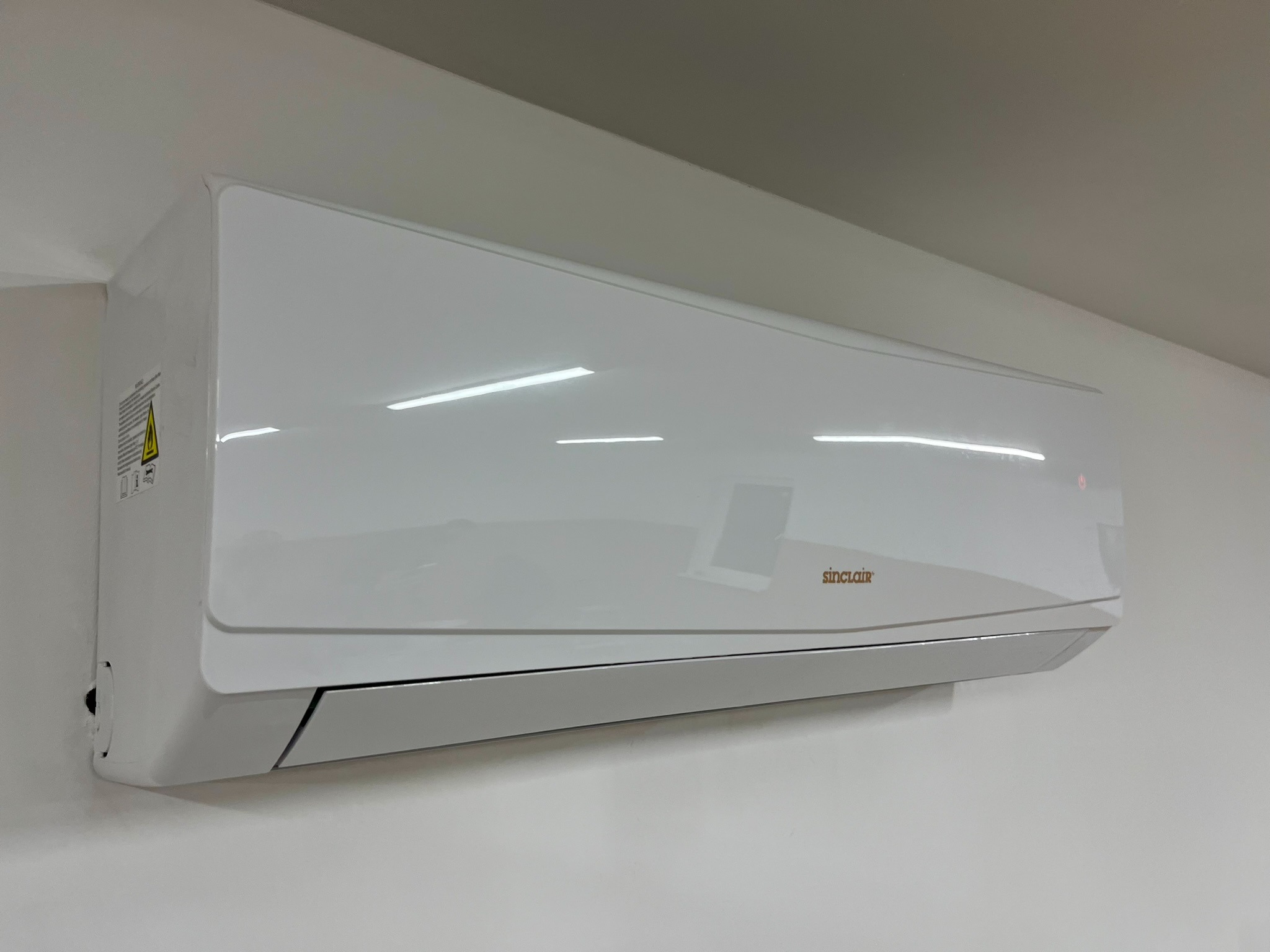 Biała jednostka klimatyzacji Sinclair zamontowana na jasnej ścianie, widoczna dioda sygnalizacyjna i naklejka ostrzegawcza po lewej stronie.