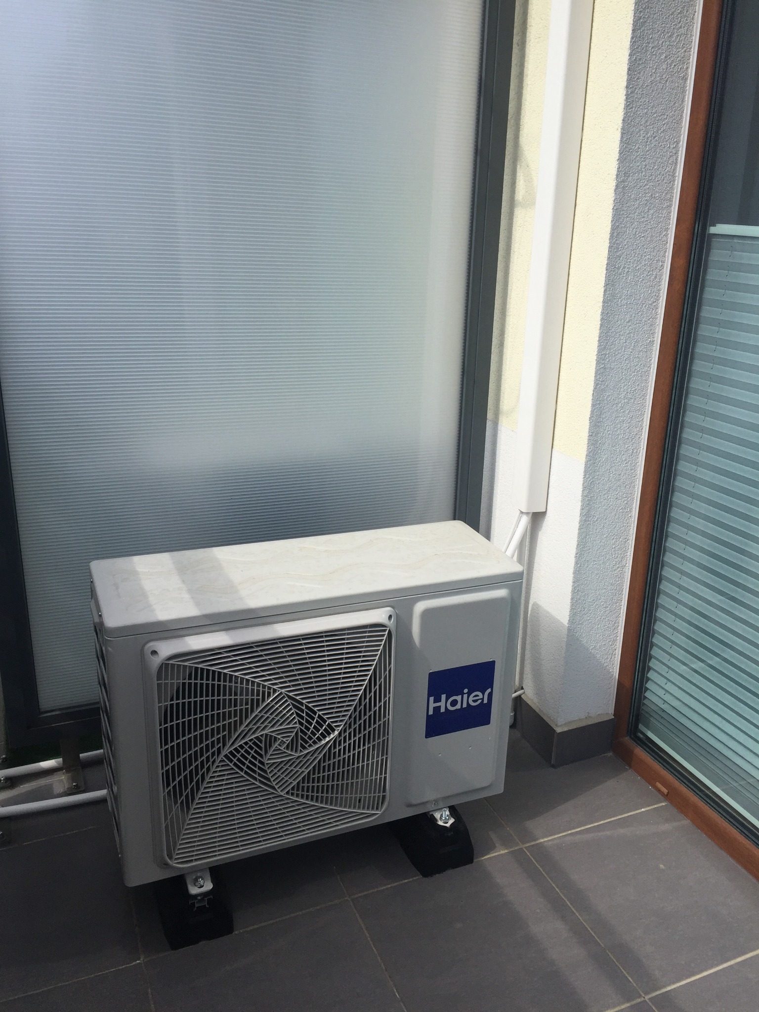 Kompaktowy klimatyzator Haier na balkonie, ustawiony na czarnych podkładkach antywibracyjnych, widoczne rury odprowadzające skropliny, tło stanowią przesuwne drzwi balkonowe z żaluzjami.