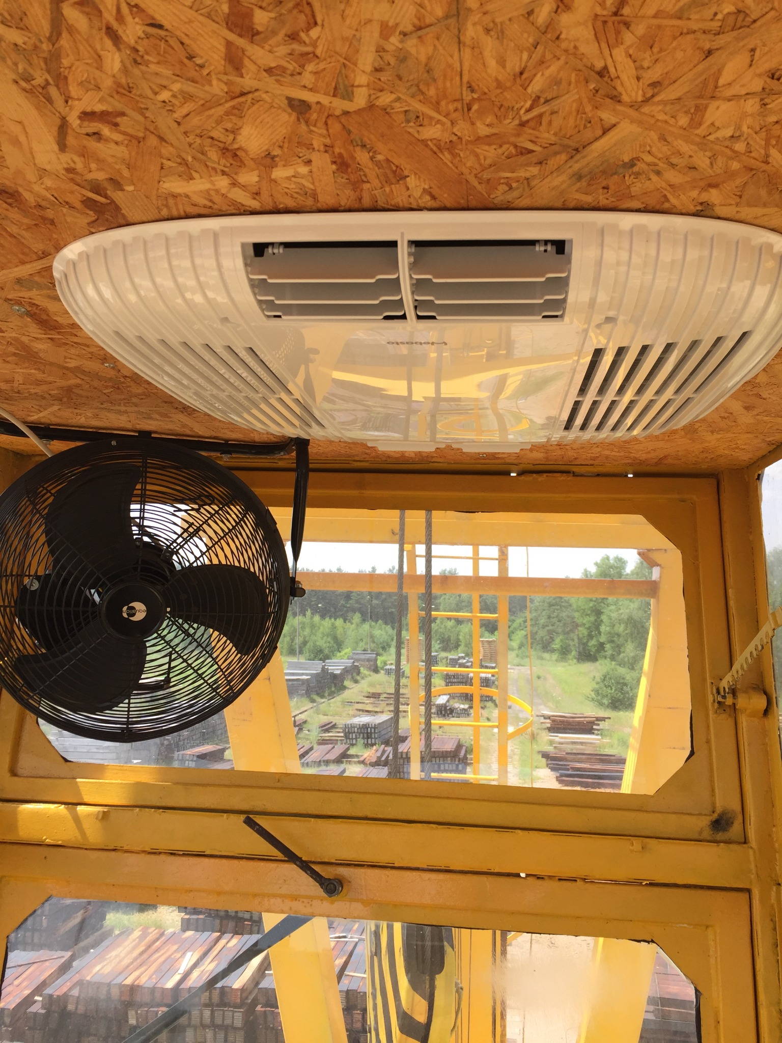 Wnętrze kabiny maszyny budowlanej w Bielsku Podlaskim z zamontowaną klimatyzacją sufitową i wentylatorem, widok na teren składowania materiałów budowlanych.