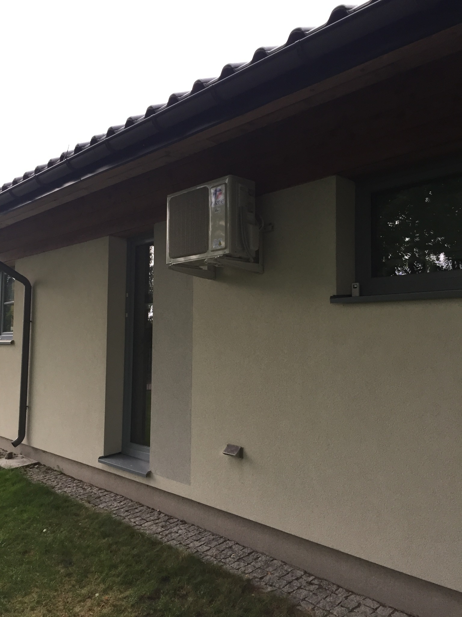 Zewnętrzna jednostka klimatyzacji zamontowana na elewacji domu jednorodzinnego, widoczna z boku, z fragmentem okna i dachu w tle.