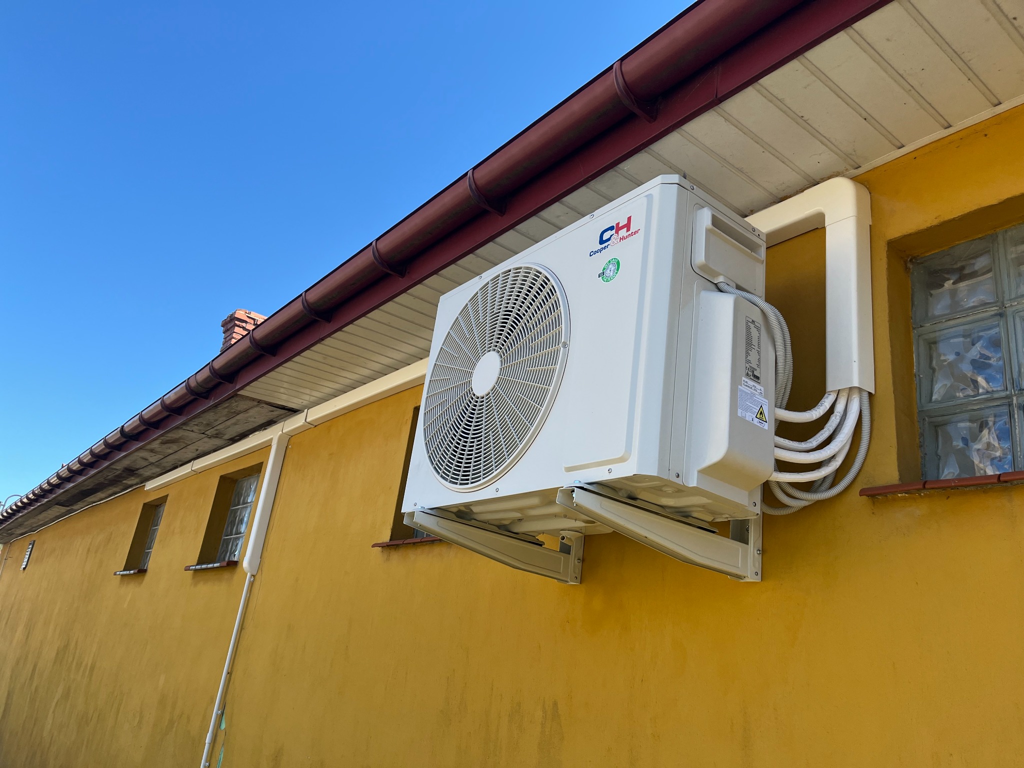 Zewnętrzna jednostka klimatyzacji Cooper&Hunter zamontowana na żółtej ścianie budynku, widoczne rury i kable w osłonie, okno z luksferami w tle.