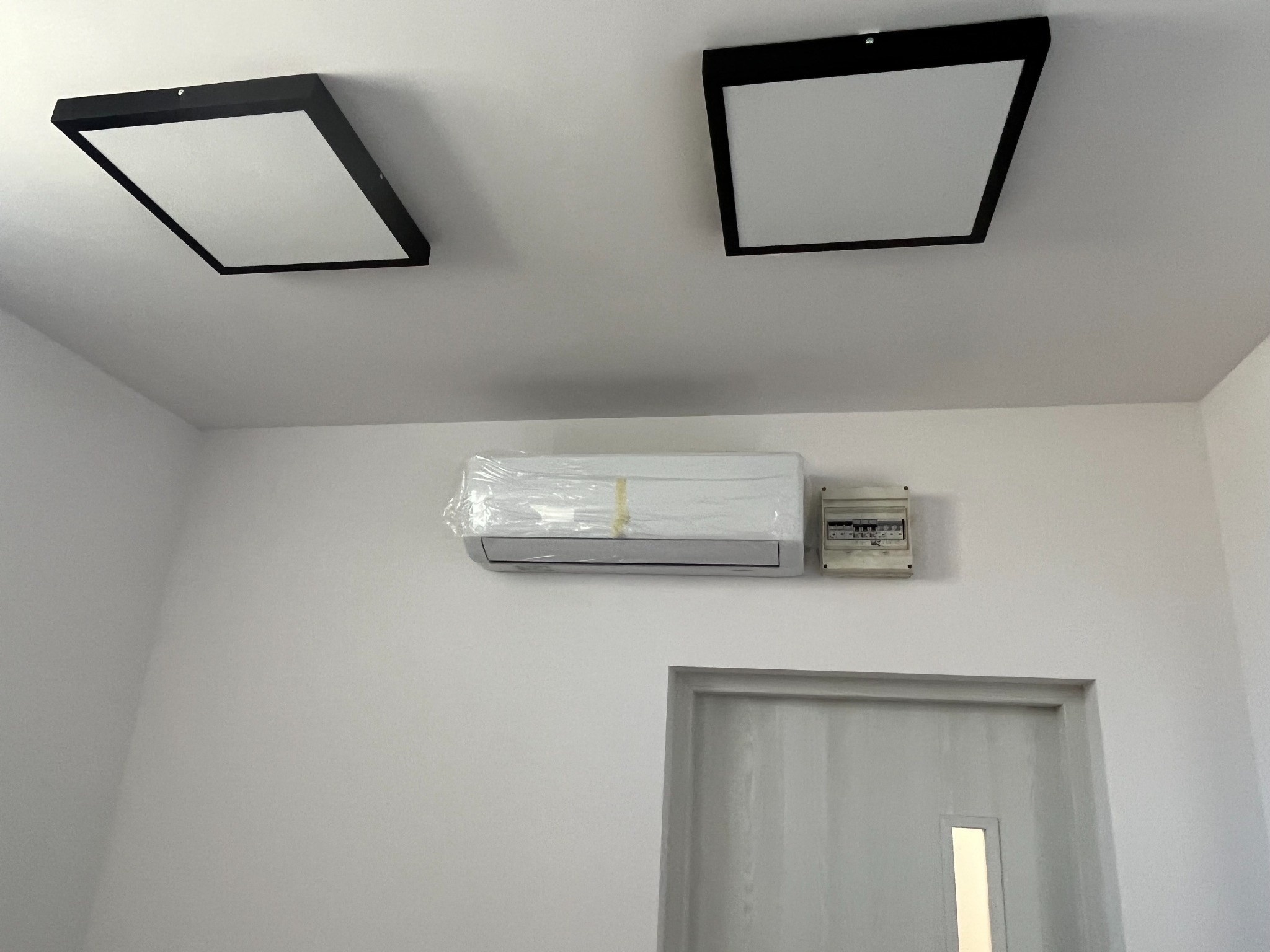 Białe pomieszczenie z zamontowaną klimatyzacją, jeszcze w folii ochronnej, obok skrzynka elektryczna i nad drzwiami, na suficie dwa kwadratowe panele LED z czarną ramką.