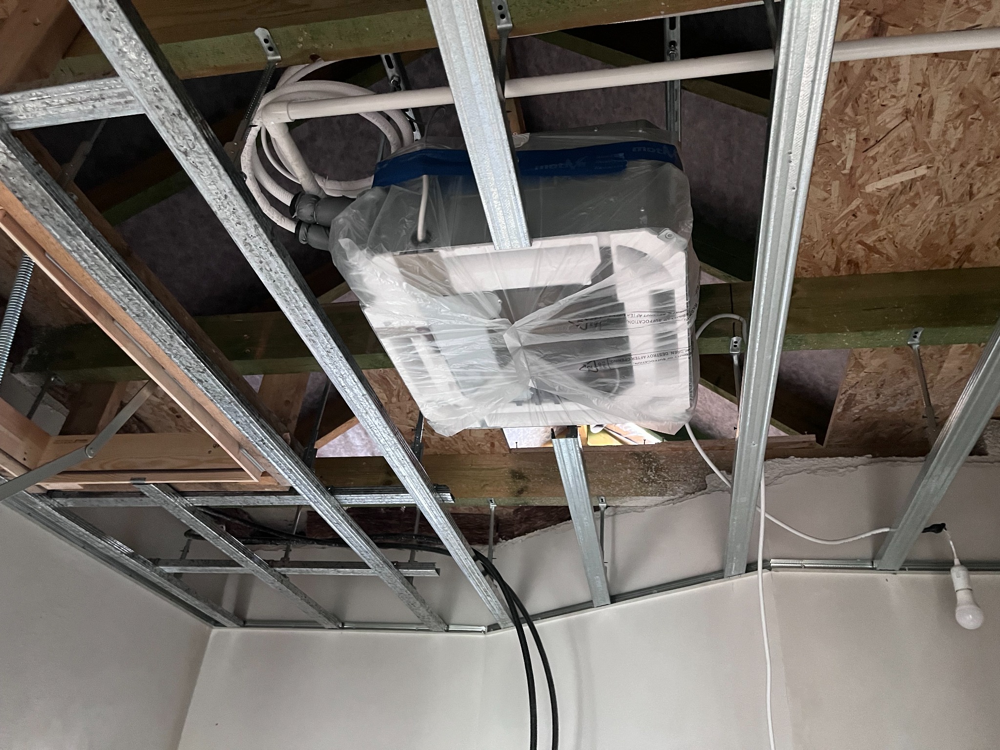 Montaż klimatyzatora kasetonowego pod sufitem podwieszanym, widoczne rury i przewody, zabezpieczony folią ochronną przed zabrudzeniem podczas prac wykończeniowych.