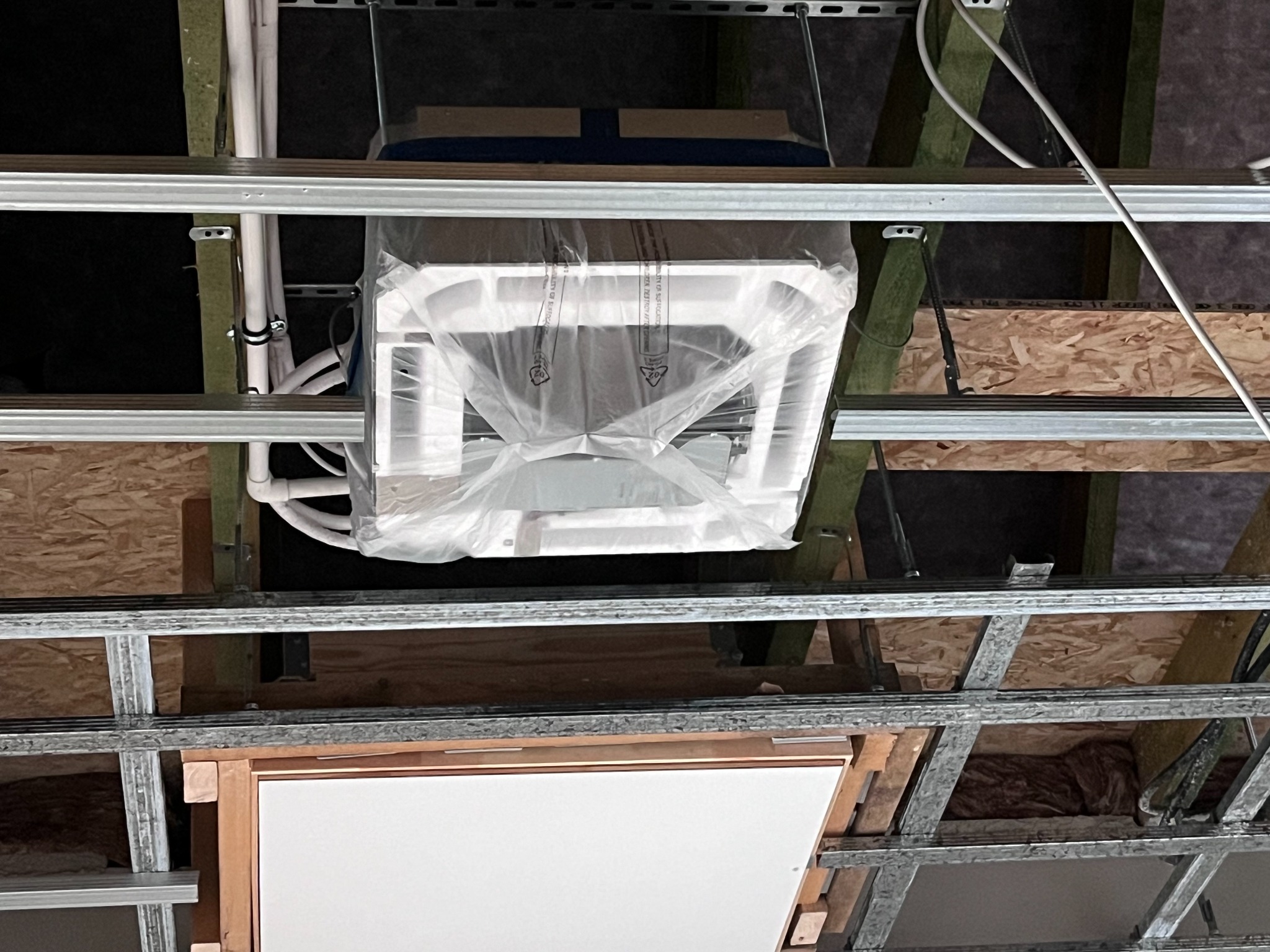 Nierozpakowana jednostka wewnętrzna klimatyzacji zamontowana w przestrzeni pod sufitem podwieszanym, widoczne rury instalacyjne i elementy konstrukcyjne sufitu.