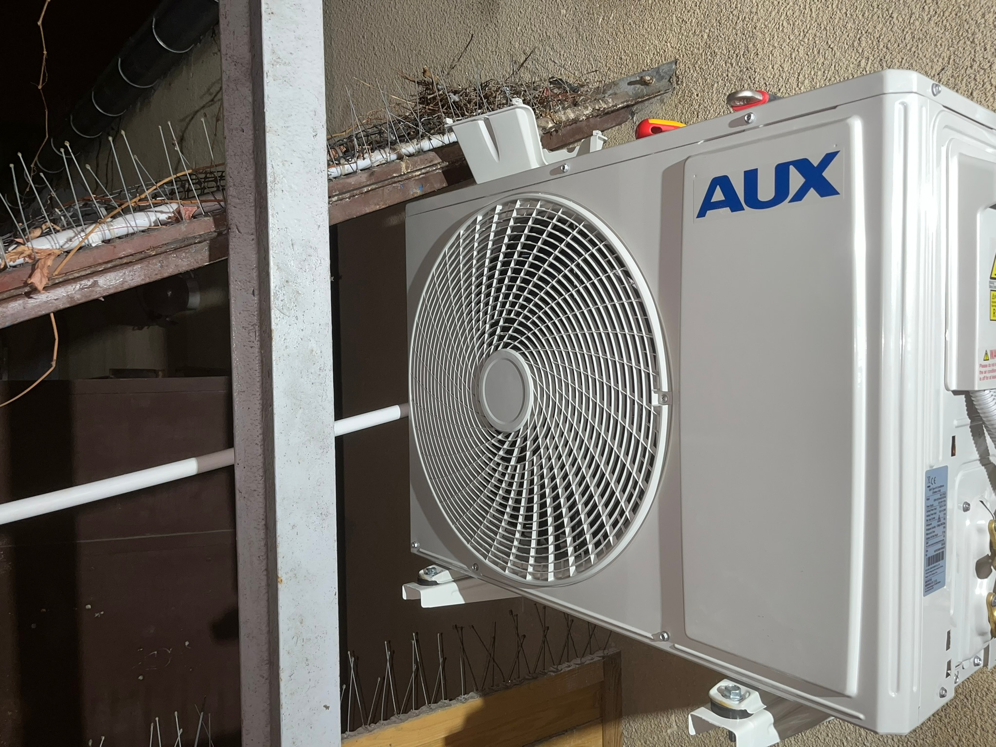 Zewnętrzna jednostka klimatyzacji AUX zamontowana na ścianie budynku z kolcami przeciw ptakom na dachu w tle.