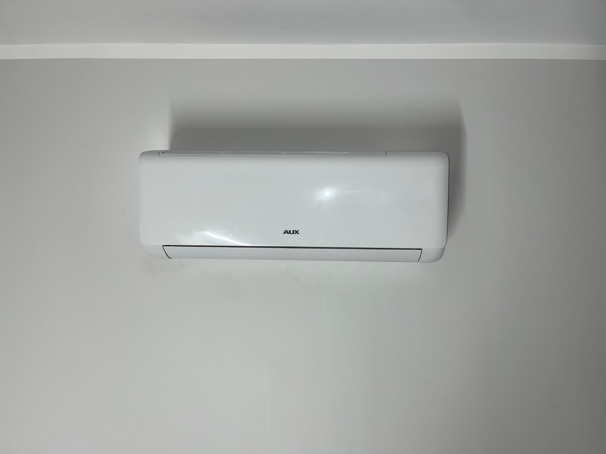 Biały klimatyzator marki AUX zamontowany na jasnoszarej ścianie w pomieszczeniu z białym sufitem.