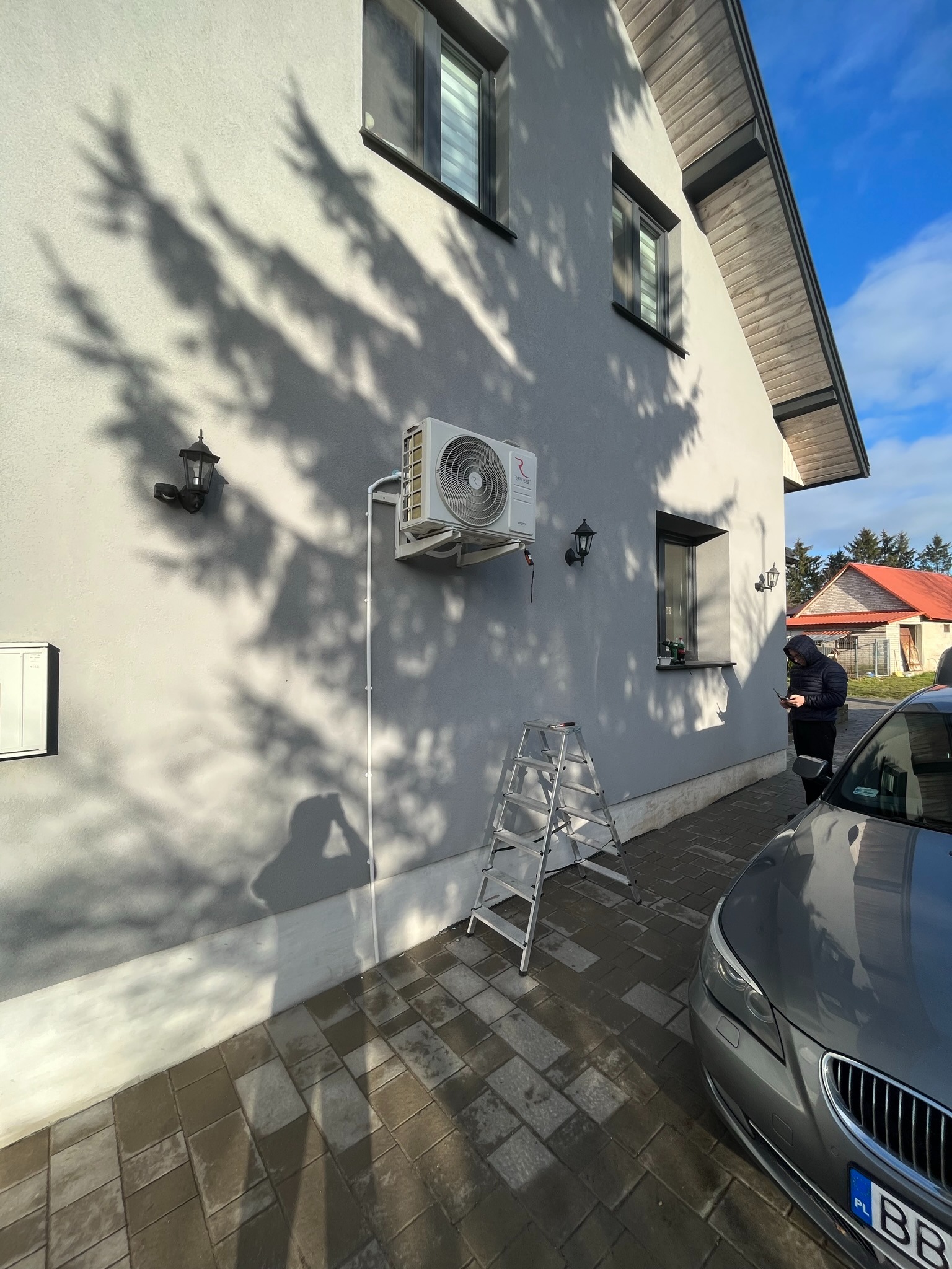 Biała jednostka zewnętrzna klimatyzacji zamontowana na szarej ścianie domu z widocznymi cieniami drzew, drabina aluminiowa oparta o ścianę, mężczyzna w kurtce sprawdza telefon, szary samochód...