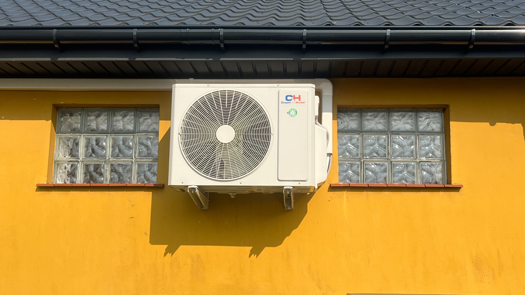 Biała jednostka zewnętrzna klimatyzacji Cooper&Hunter zamontowana na żółtej ścianie budynku, między dwoma oknami z luksferów, pod czarnym dachem z rynną.