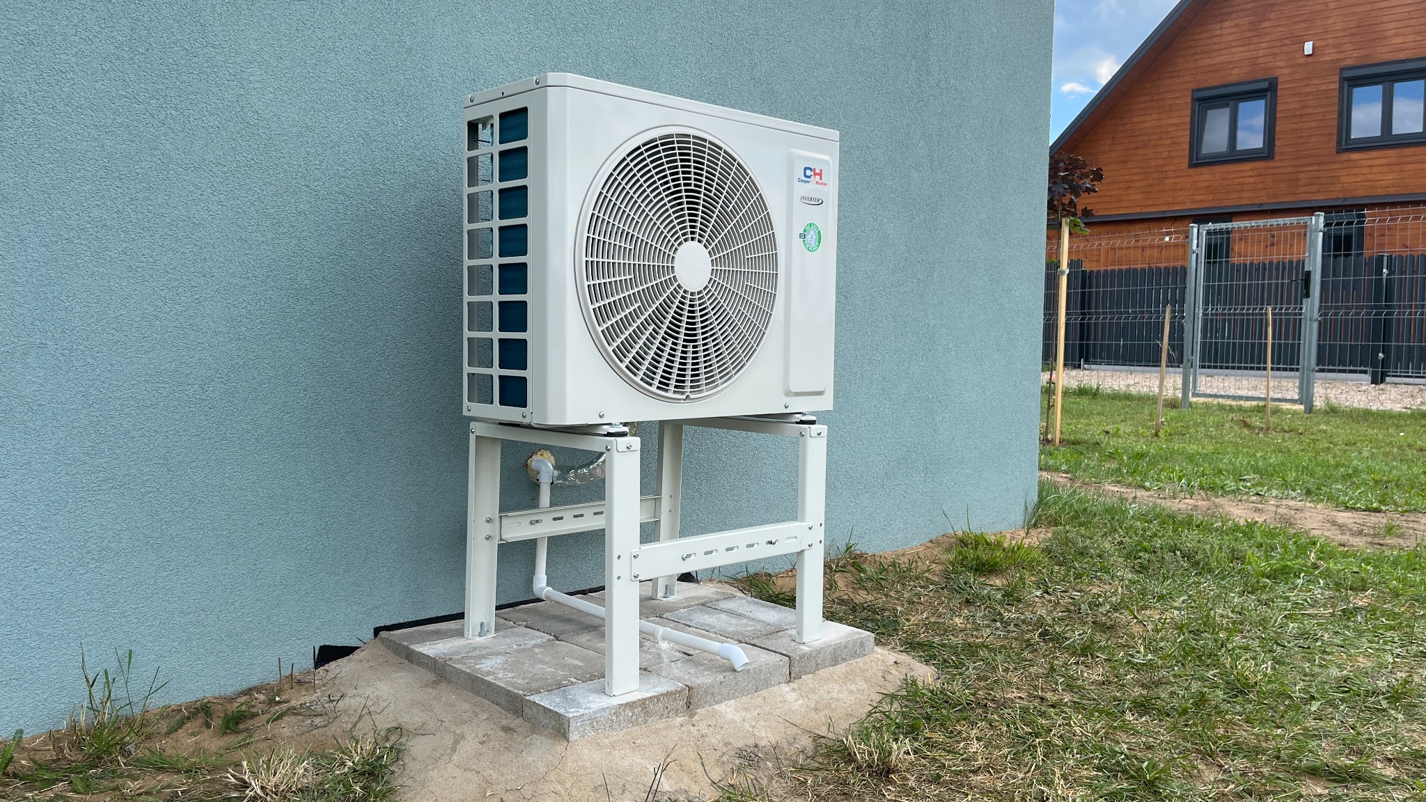 Biała jednostka zewnętrzna klimatyzacji marki Cooper&Hunter zamontowana na metalowym stelażu przy ścianie domu o błękitnym odcieniu, widoczny trawnik i fragment drewnianego domu w tle.