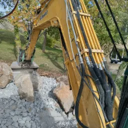 Żółta koparka Yanmar z widocznymi przewodami hydraulicznymi i ramieniem, pracująca na terenie z kamieniami i gruzem, widok z kabiny operatora na tle drzew i budynku.