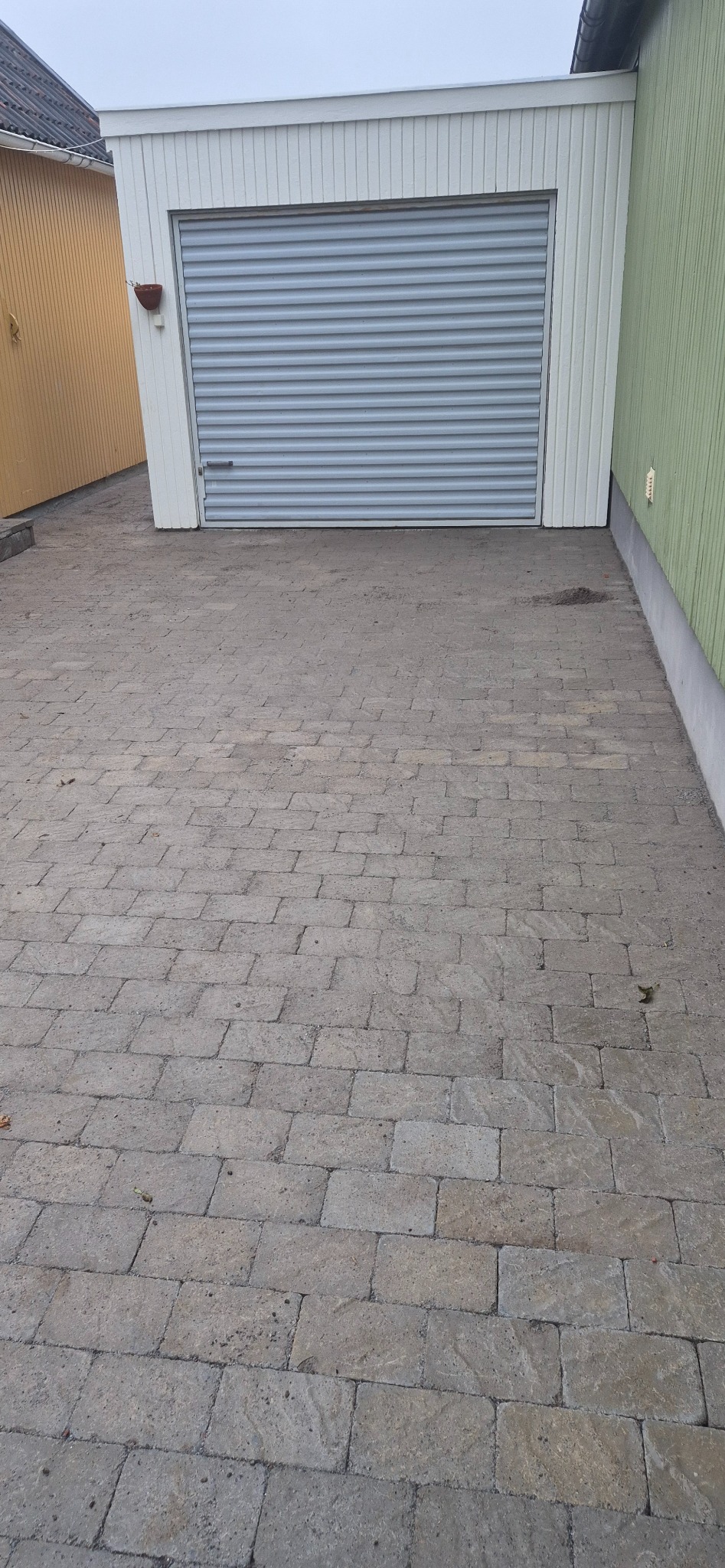 Szara kostka brukowa ułożona przed garażem z szarą bramą rolowaną. Widoczne sąsiednie budynki o żółtej i zielonej elewacji. Pochmurne niebo.