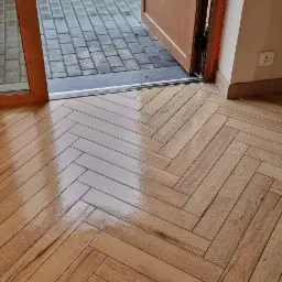 Parkiet ułożony we wzór jodełki francuskiej, widok z perspektywy otwartych drzwi na zewnątrz, gdzie widać mokry bruk i rower.