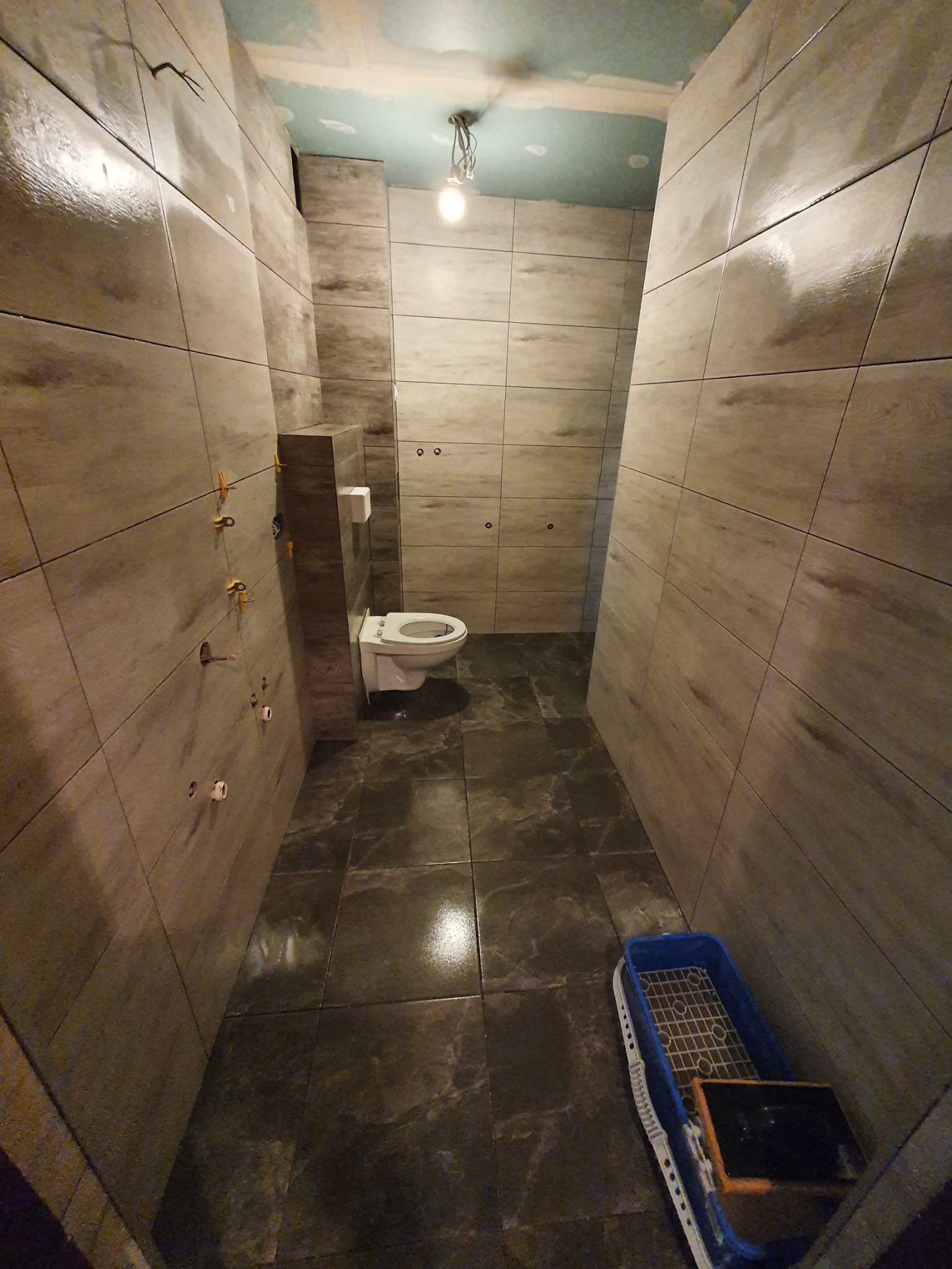 Wąska łazienka w trakcie prac wykończeniowych, widoczne płytki imitujące drewno na ścianach, ciemne płytki podłogowe, podwieszana toaleta, narzędzia do układania płytek oraz niezakończone prace...