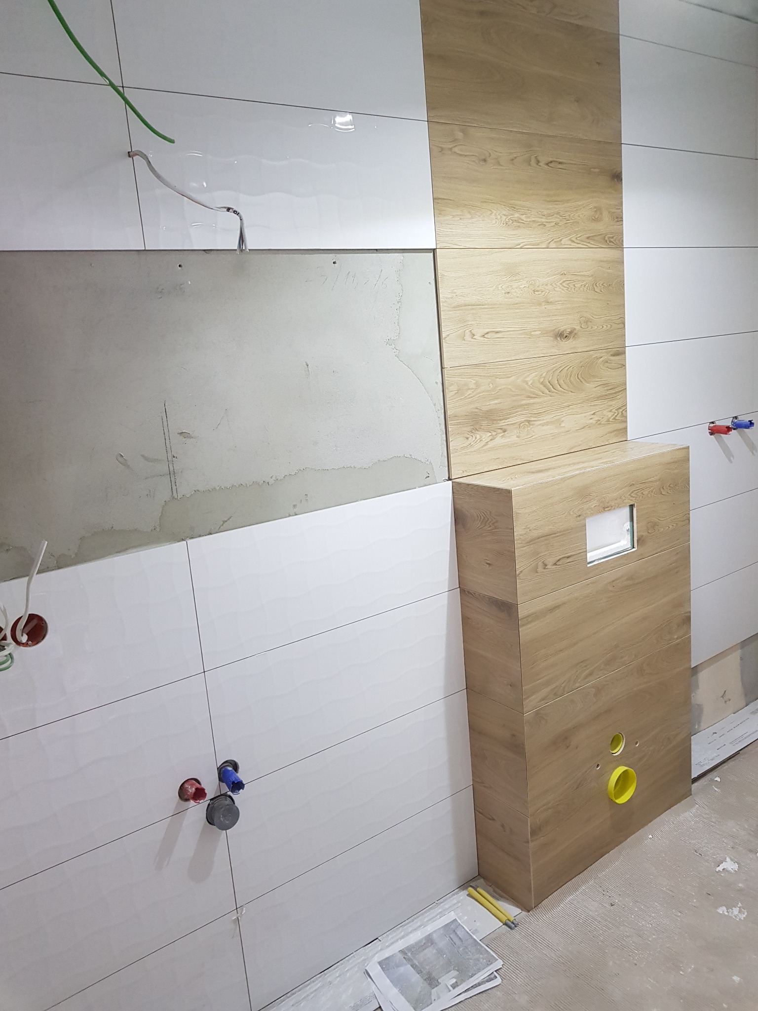 Łazienka w trakcie remontu: białe płytki ścienne z subtelną falą, fragment ściany z surowym betonem, drewnopodobne płytki na zabudowie stelaża WC z widocznym otworem rewizyjnym i przyłączami...