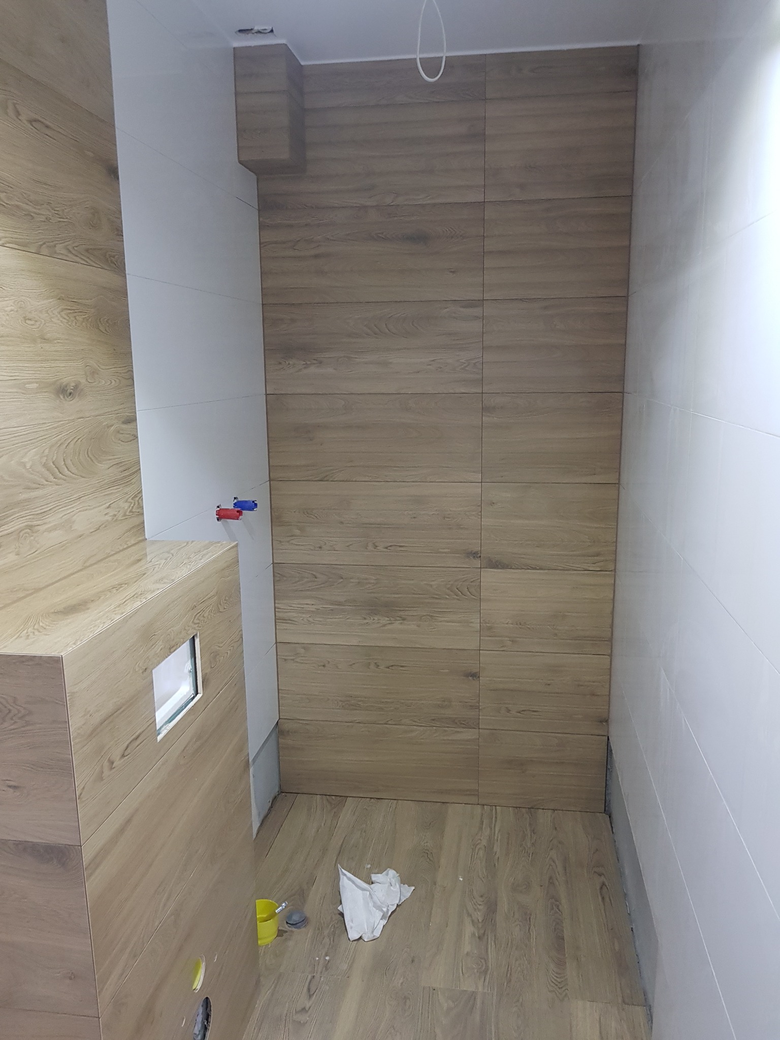 Nowoczesna łazienka z płytkami imitującymi drewno na ścianach i podłodze, białe płytki na pozostałych ścianach, widoczne przyłącza wodne i elementy instalacji, nieukończone prace wykończeniowe.