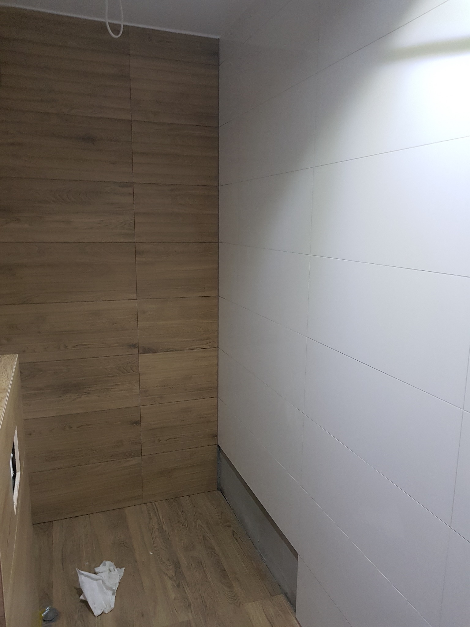 Fragment łazienki z wyłożonymi płytkami imitującymi drewno na podłodze i jednej ścianie, oraz białymi płytkami na drugiej ścianie, z widocznym niedokończonym wykończeniem przy podłodze i leżącą...