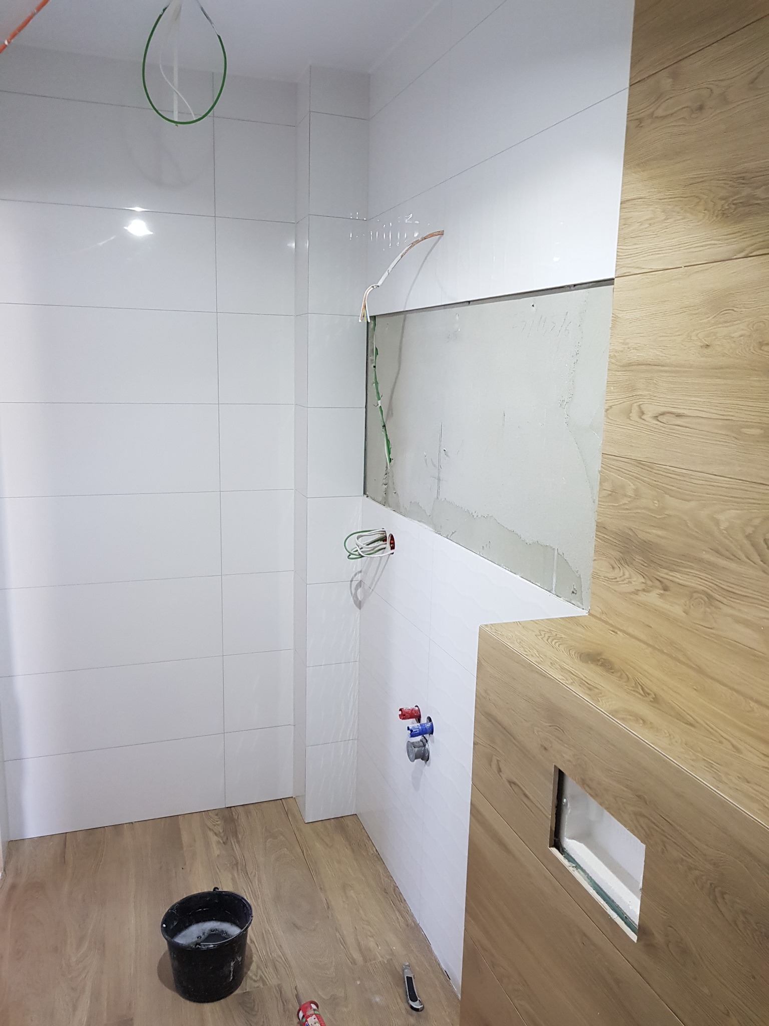 Wnętrze łazienki w trakcie prac: białe płytki ścienne, fragment ściany bez wykończenia, drewnopodobna obudowa, wystające kable, wiadro z wodą i narzędzia na podłodze.