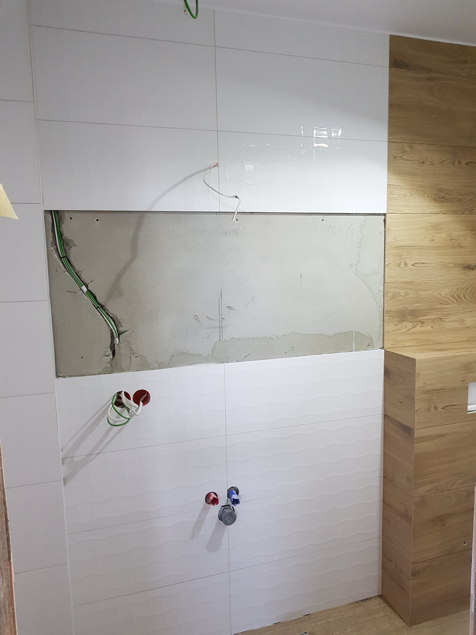 Ściana łazienki w trakcie wykańczania z widocznymi przewodami elektrycznymi i rurami wodnymi, częściowo pokryta białymi płytkami z delikatnym wzorem fal, obok fragment ściany wyłożony panelami...