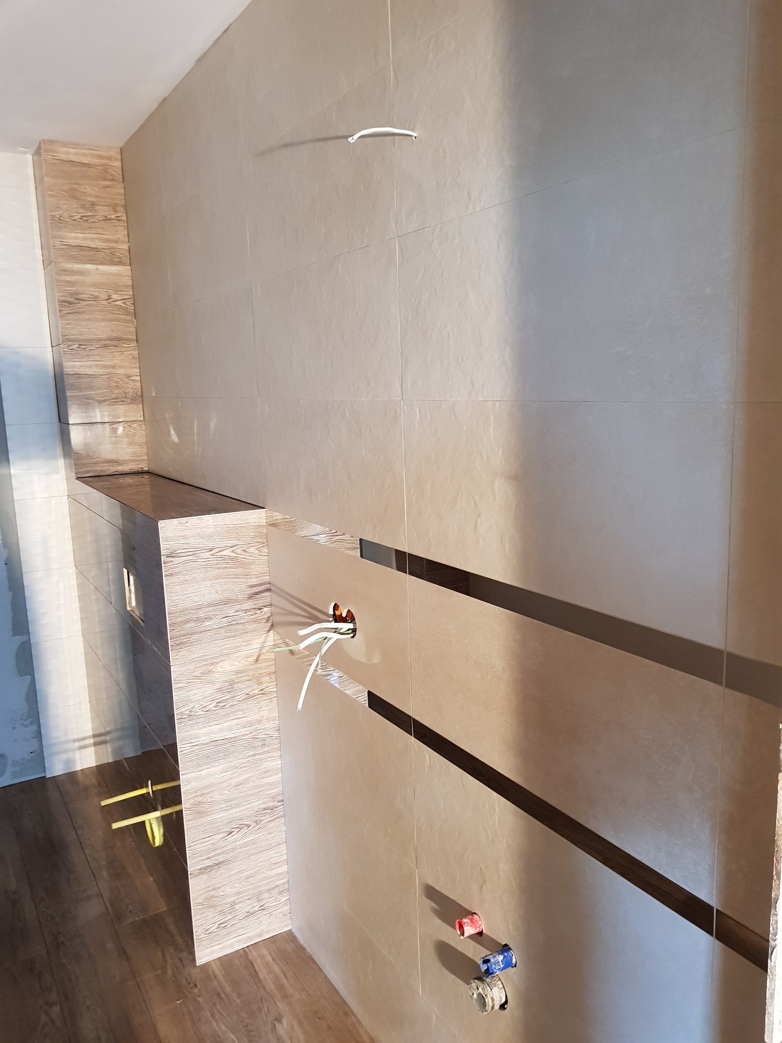 Łazienka w trakcie remontu: beżowe płytki ścienne z ciemnobrązowymi pasami dekoracyjnymi, zabudowa geberitu z płytkami imitującymi drewno, wystające przewody elektryczne i rury wodne.