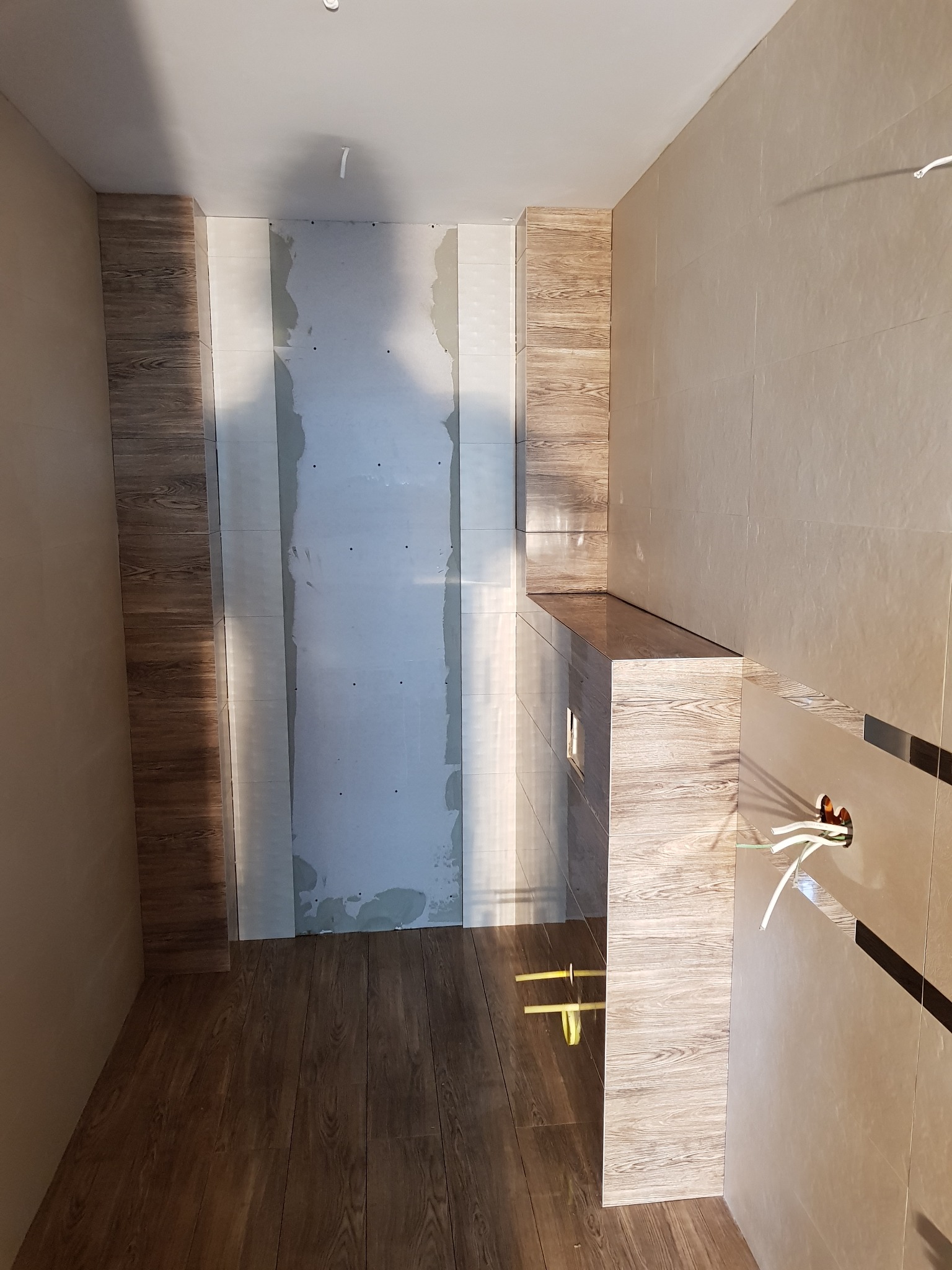 Wnętrze łazienki w trakcie prac wykończeniowych: ułożona podłoga z paneli imitujących drewno, częściowo wyłożone ściany beżowymi płytkami, fragment ściany przygotowany pod gładzie, wystające kable...