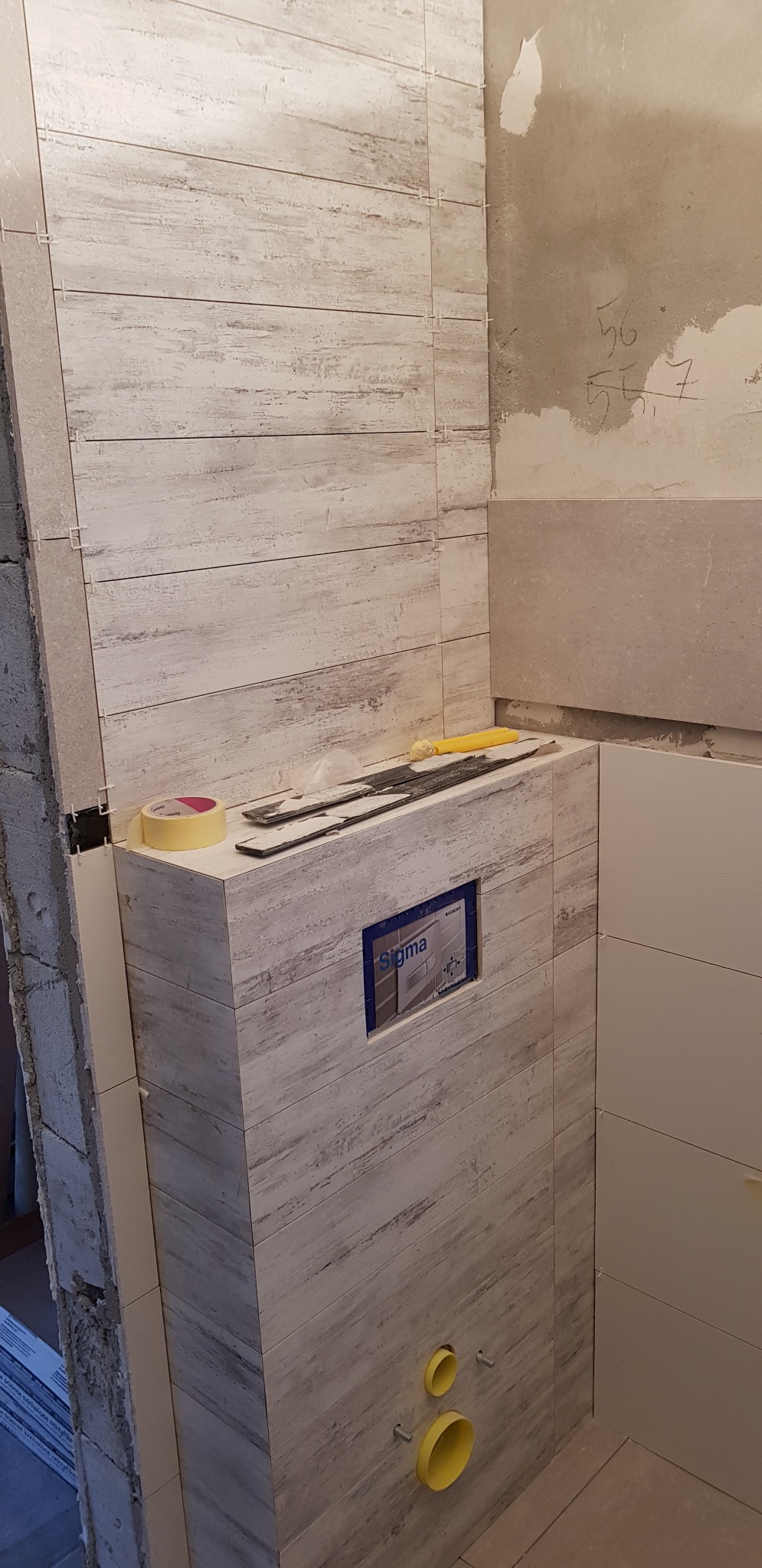 Ściana łazienki w trakcie wykańczania, z widocznymi płytkami imitującymi drewno, żółtą taśmą malarską, narzędziami i elementami instalacji sanitarnych.