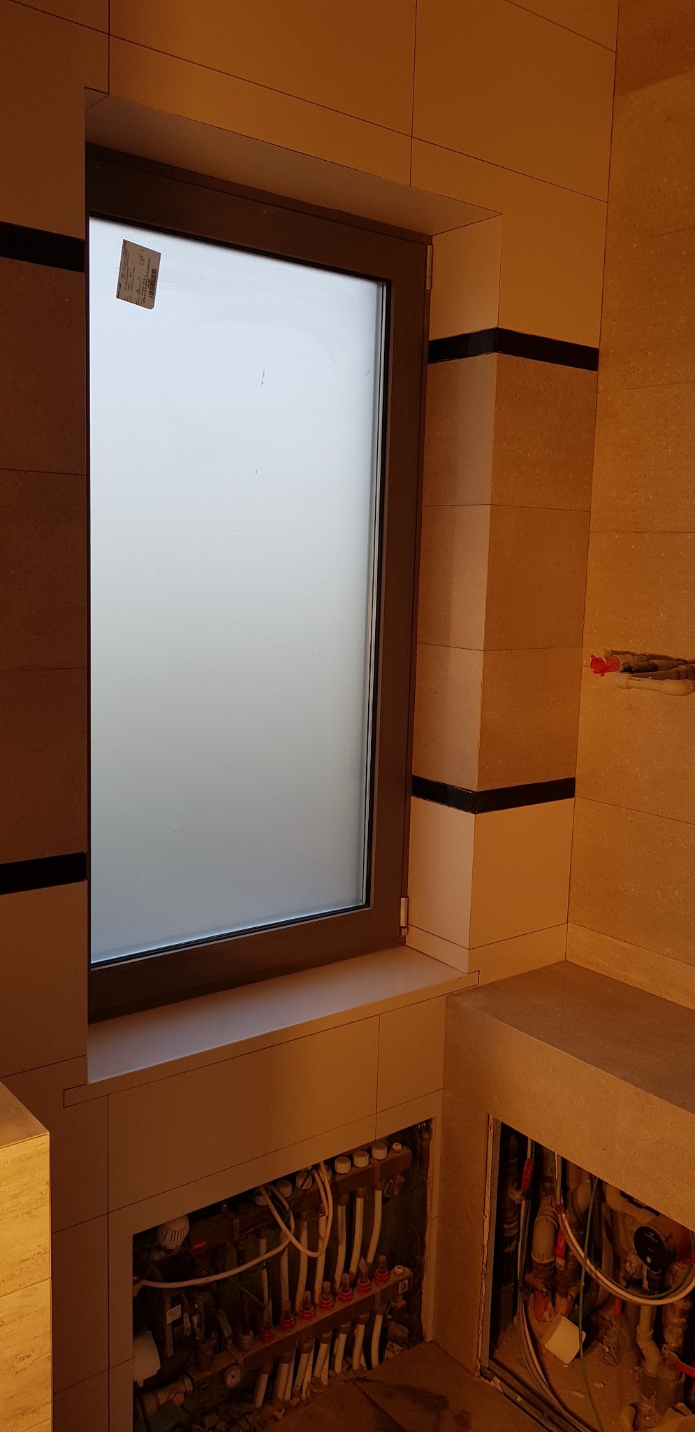 Łazienka w trakcie wykańczania w Perzowie: widok na okno z naklejką, fragment zabudowy z beżowymi płytkami z czarnymi pasami oraz otwarte przestrzenie z instalacją grzewczą pod podłogą.