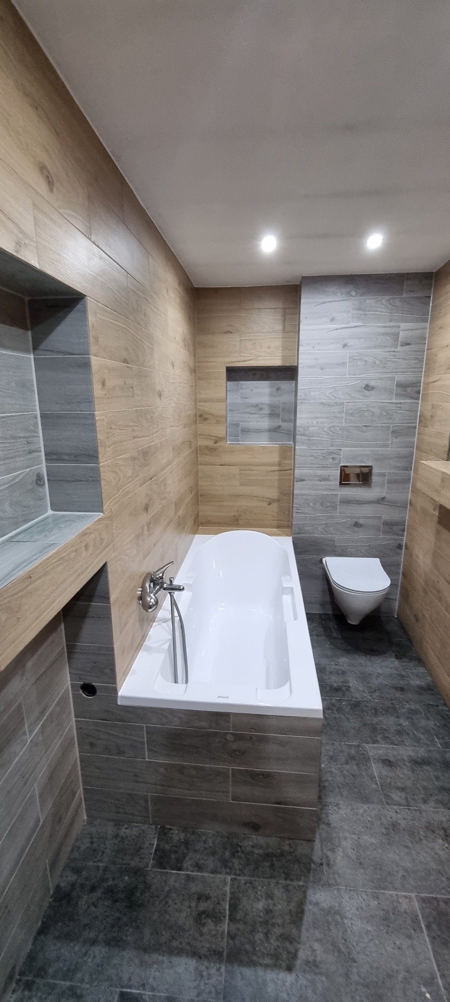 Nowoczesna łazienka z wanną obudowaną płytkami imitującymi drewno, szare płytki na ścianach i podłodze, oraz wisząca toaleta.