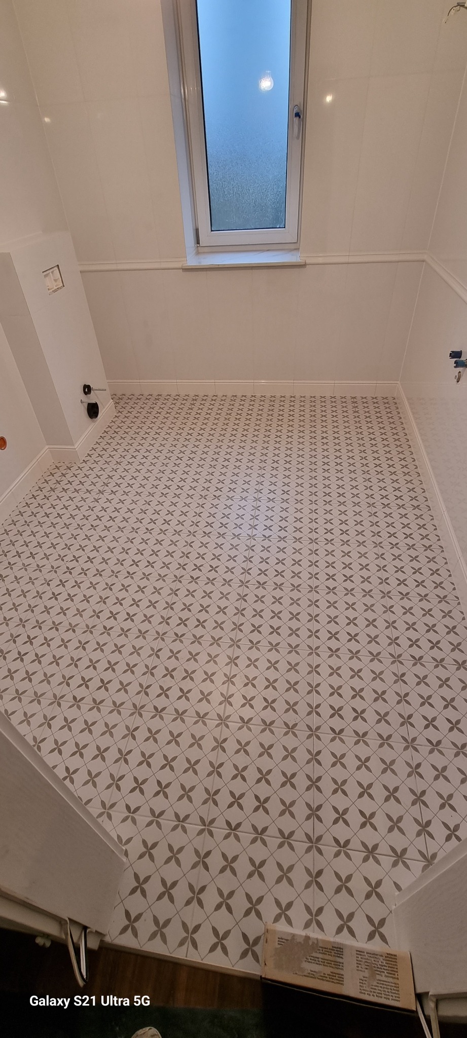 Wykończona łazienka z białymi płytkami ściennymi i podłogą w geometryczny wzór kwiatowy, widok z góry na okno dachowe.