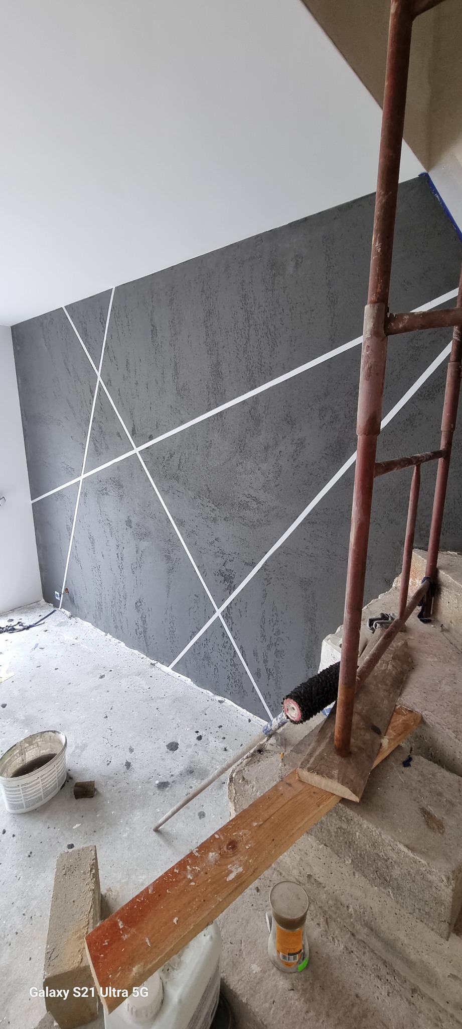 Ściana pokryta szarą, strukturalną farbą z geometrycznym wzorem z białych pasów, widoczne rusztowanie i narzędzia malarskie w surowym wnętrzu w trakcie prac wykończeniowych.