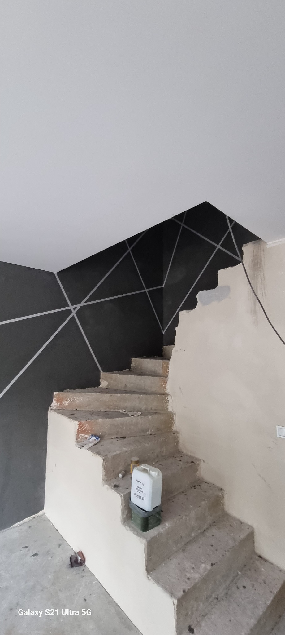 Schody w trakcie prac wykończeniowych w Perzowie, z pomalowaną na czarno ścianą z geometrycznym wzorem i widocznym pojemnikiem z masą szpachlową.