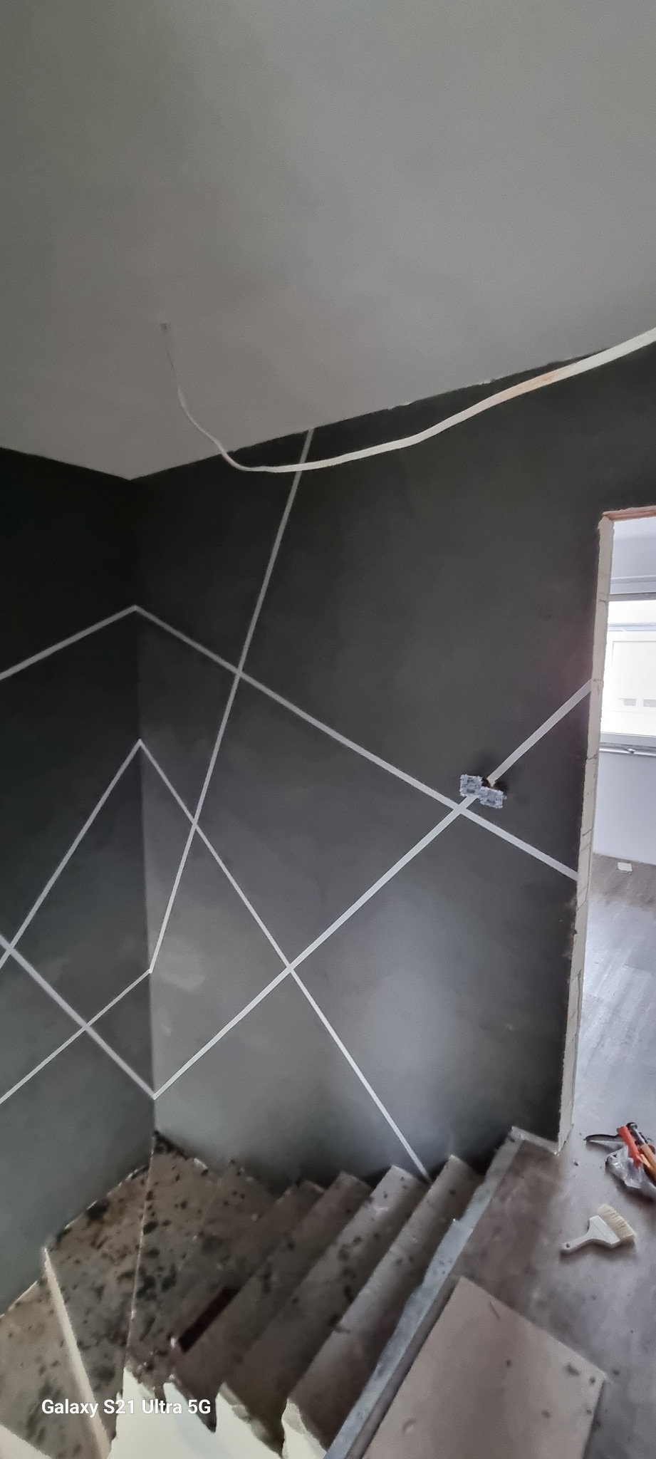 Ściana pomalowana na szaro z geometrycznym wzorem z białej taśmy malarskiej, widoczne schody w stanie surowym i wystające przewody elektryczne.