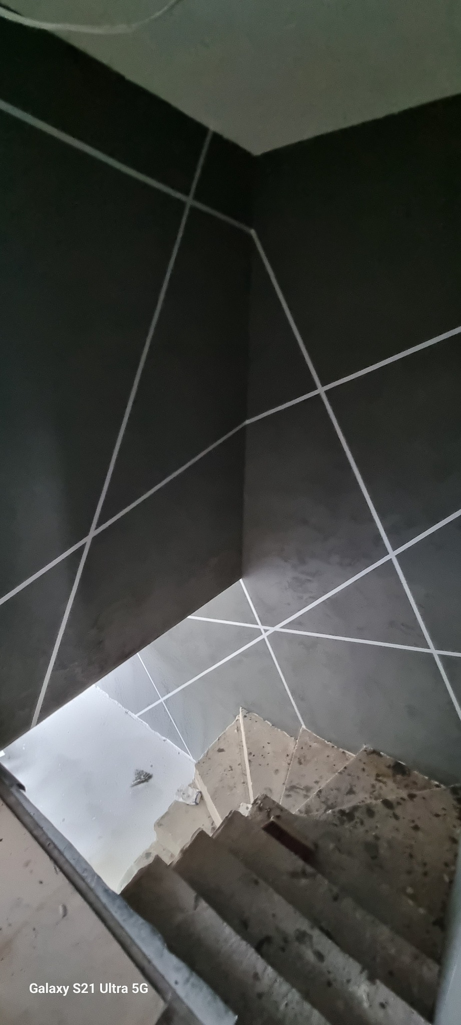 Szare ściany w klatce schodowej z dekoracyjnymi białymi paskami tworzącymi geometryczny wzór, widoczne schody w trakcie prac wykończeniowych.