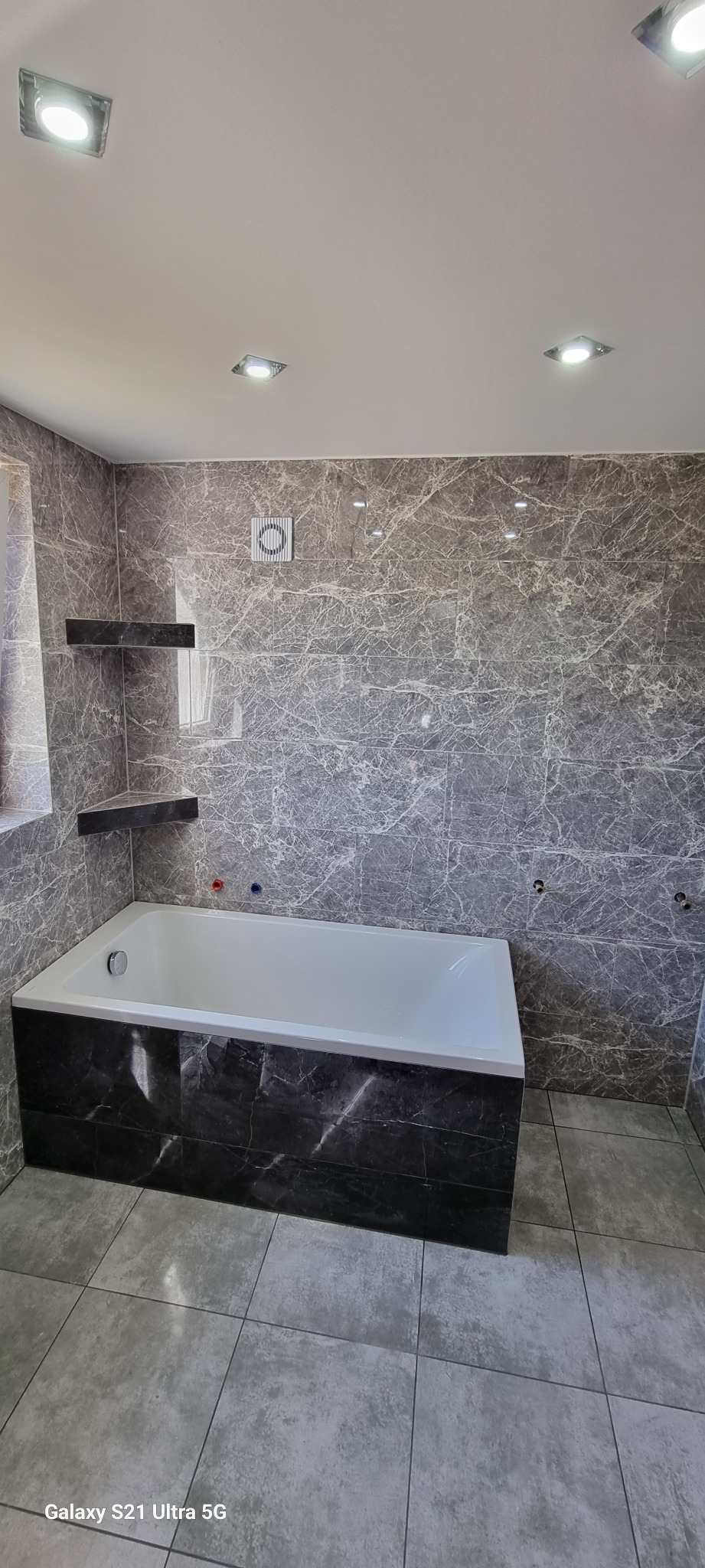 Wykończona łazienka z wanną obudowaną ciemnymi płytkami imitującymi marmur, ściany wyłożone szarymi płytkami z efektem marmuru, dwie półki narożne z ciemnego marmuru, wentylator na ścianie...