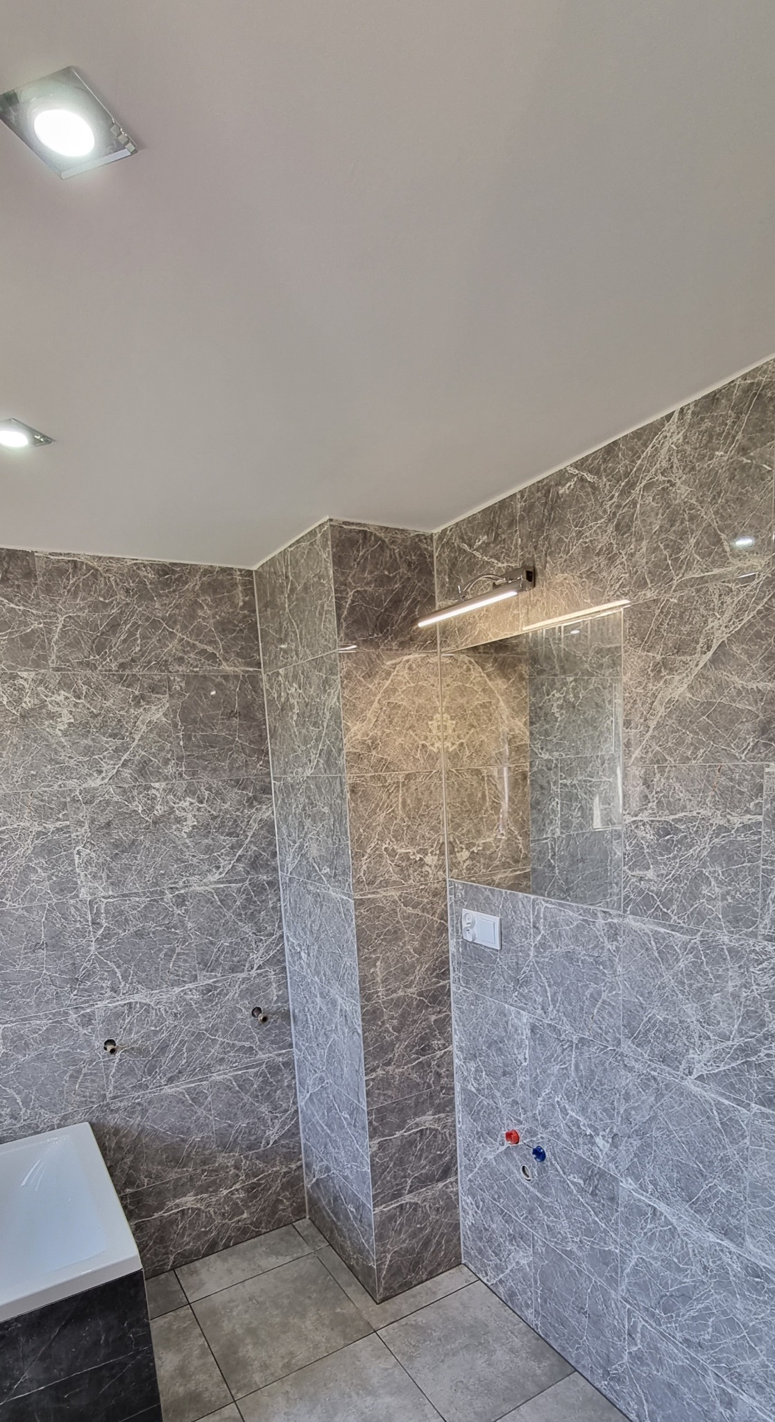 Wykończona łazienka z szarymi płytkami imitującymi marmur na ścianach i podłodze, widoczne przyłącza wodne, lustro z oświetleniem LED i fragment wanny po lewej stronie.