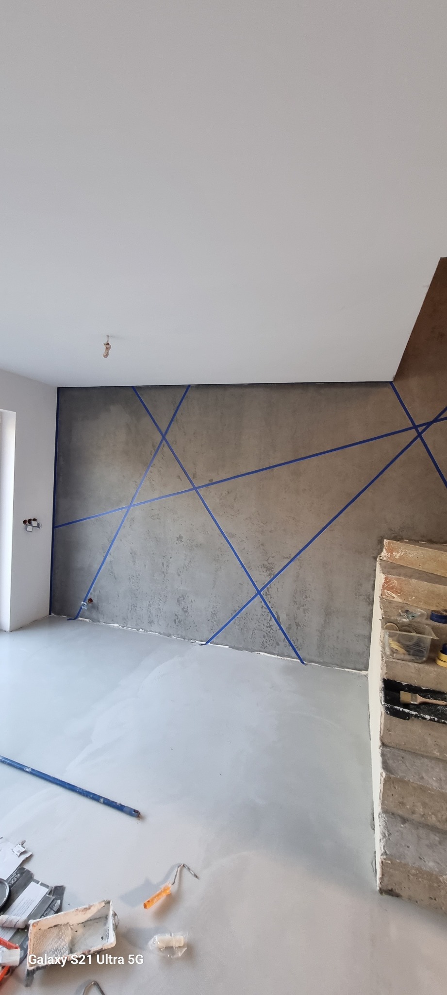 Ściana w surowym stylu z szarą fakturą i niebieskimi liniami taśmy malarskiej tworzącymi geometryczny wzór, fragment podłogi zalany wylewką samopoziomującą, narzędzia malarskie na podłodze...