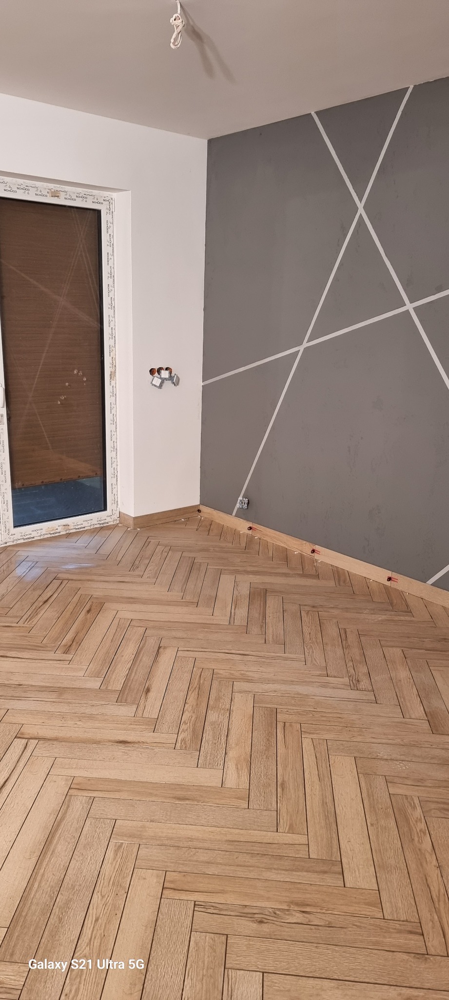 Ułożona podłoga z paneli w jodełkę, szara ściana z białymi liniami, widoczne gniazdka elektryczne bez osprzętu, listwa przypodłogowa z czerwonymi klinami dystansowymi.