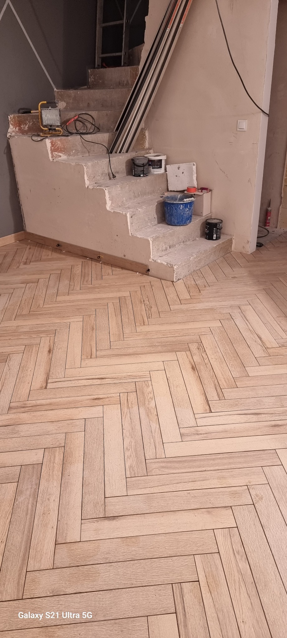 Montaż drewnopodobnych paneli podłogowych w jodełkę francuską, widoczne schody w trakcie prac przygotowawczych, z narzędziami i materiałami budowlanymi.