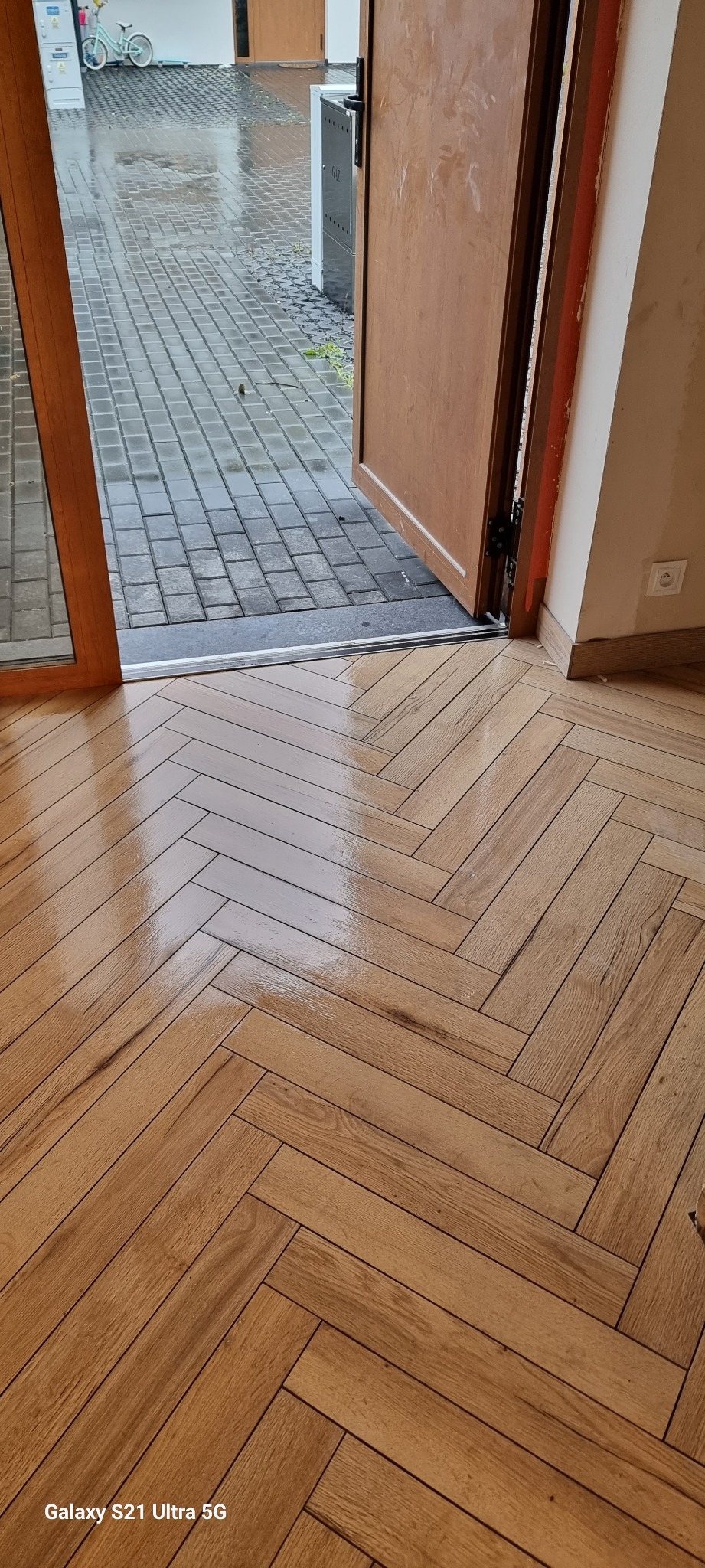 Parkiet ułożony we wzór jodełki francuskiej, widok z perspektywy otwartych drzwi na zewnątrz, gdzie widać mokry bruk i rower.