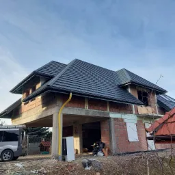 Nowy, czarny dach z dachówki ceramicznej na surowym budynku z czerwonej cegły, widoczne rynny i żółta rura spustowa, furgonetka w tle.