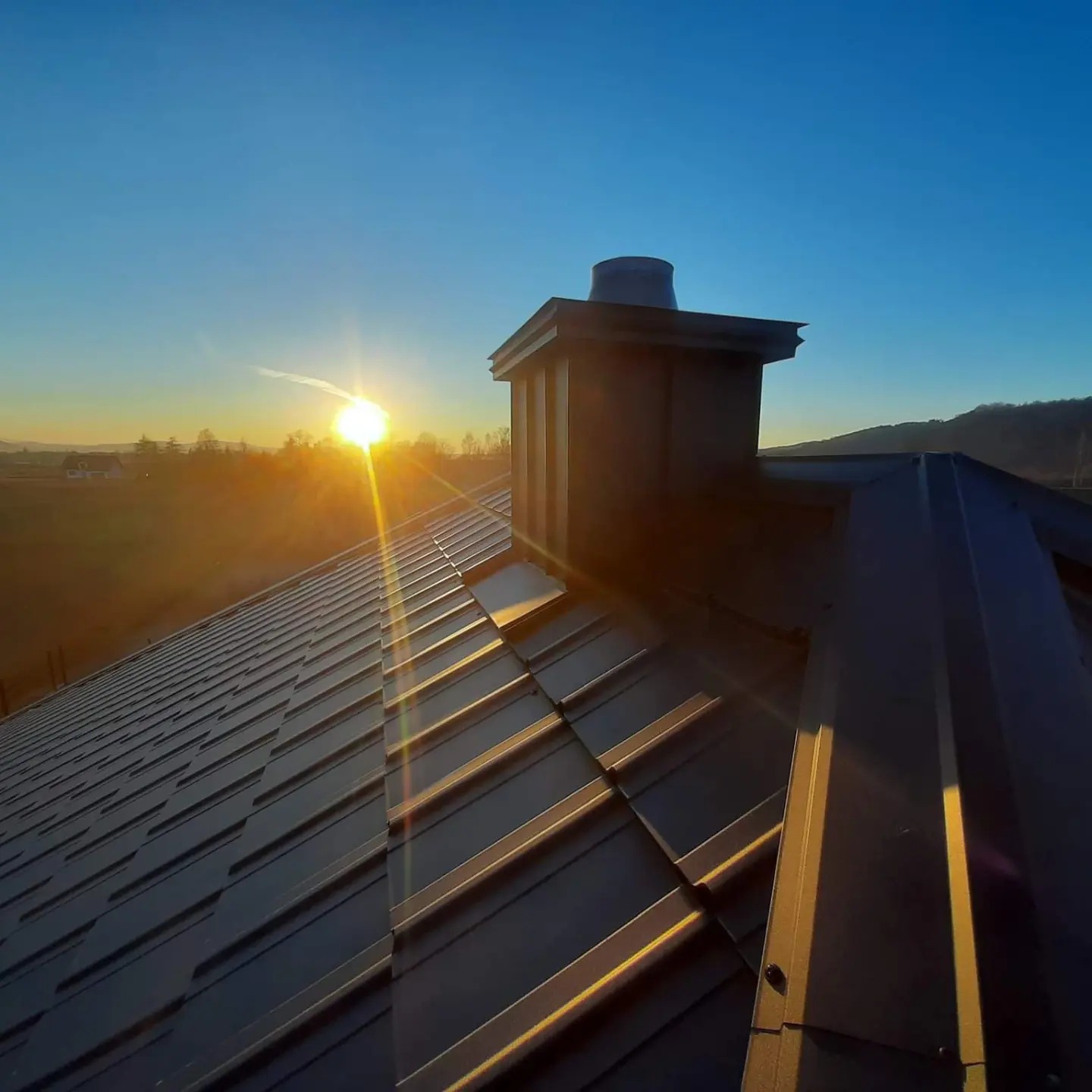 Nowoczesny, ciemny dach z blachy na tle wschodzącego słońca, widoczny komin i krajobraz wiejski w oddali.