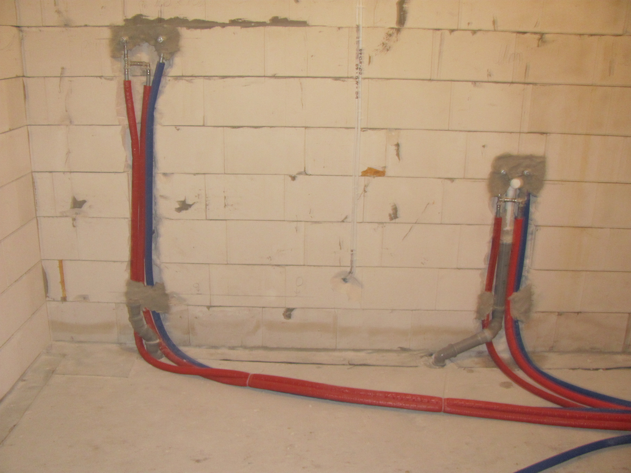 Instalacja hydrauliczna w surowym stanie: czerwone i niebieskie rury PEX wystające ze ściany z betonu komórkowego, zabezpieczone pianką montażową, prowadzone po podłodze.