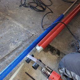 Hydro - Osa - Przygotowanie do łączenia rur PEX: niebieska i czerwona rura izolowana termicznie leżą obok zgrzewarki HYDRD-OSA na betonowej posadzce, w tle wiertarka akumulatorowa.