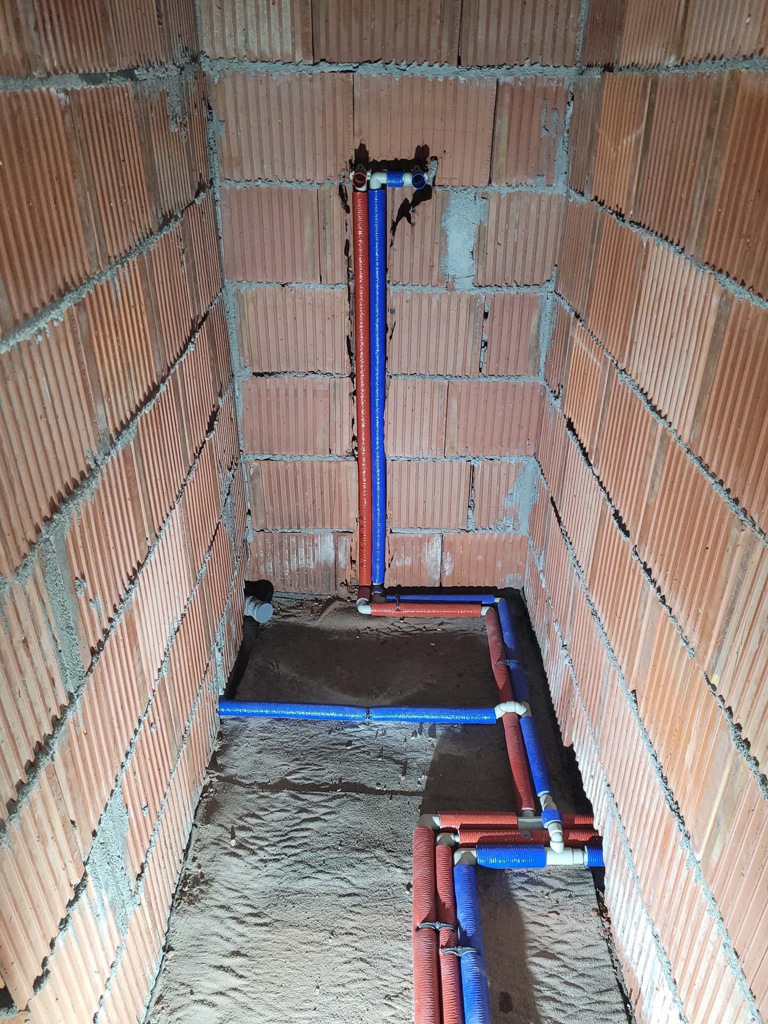 Instalacja wodna z rur PEX w kolorze niebieskim i czerwonym, ułożona na piasku w surowym pomieszczeniu z ceglanymi ścianami, widoczne złączki i kolanka.