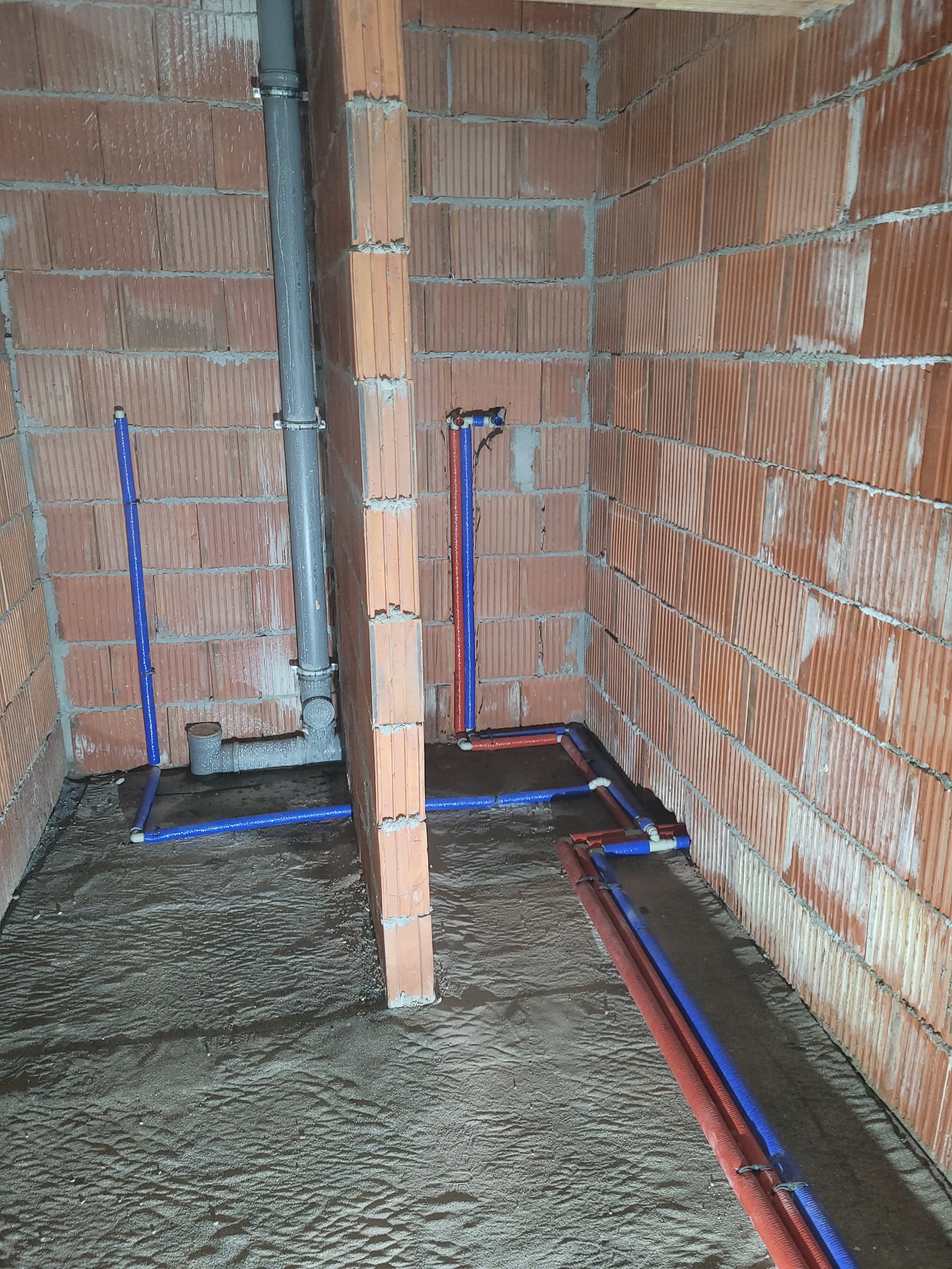 Surowy stan: instalacja wodno-kanalizacyjna w trakcie montażu, widoczne rury szarej kanalizacji i czerwono-niebieskie rury wodne, ściany z cegły dziurawki, podłoże z piasku.