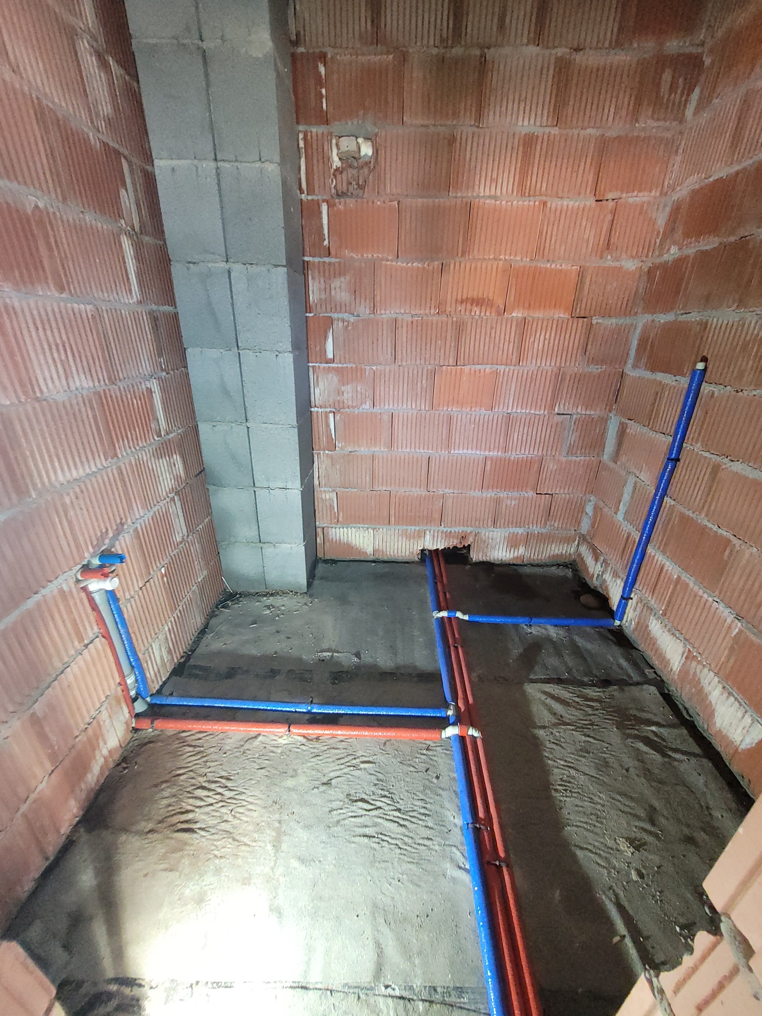 Surowy stan łazienki z widocznymi instalacjami wodnymi z rur PEX w kolorze niebieskim i czerwonym, przytwierdzonymi do ścian z czerwonej cegły i betonowego komina wentylacyjnego.