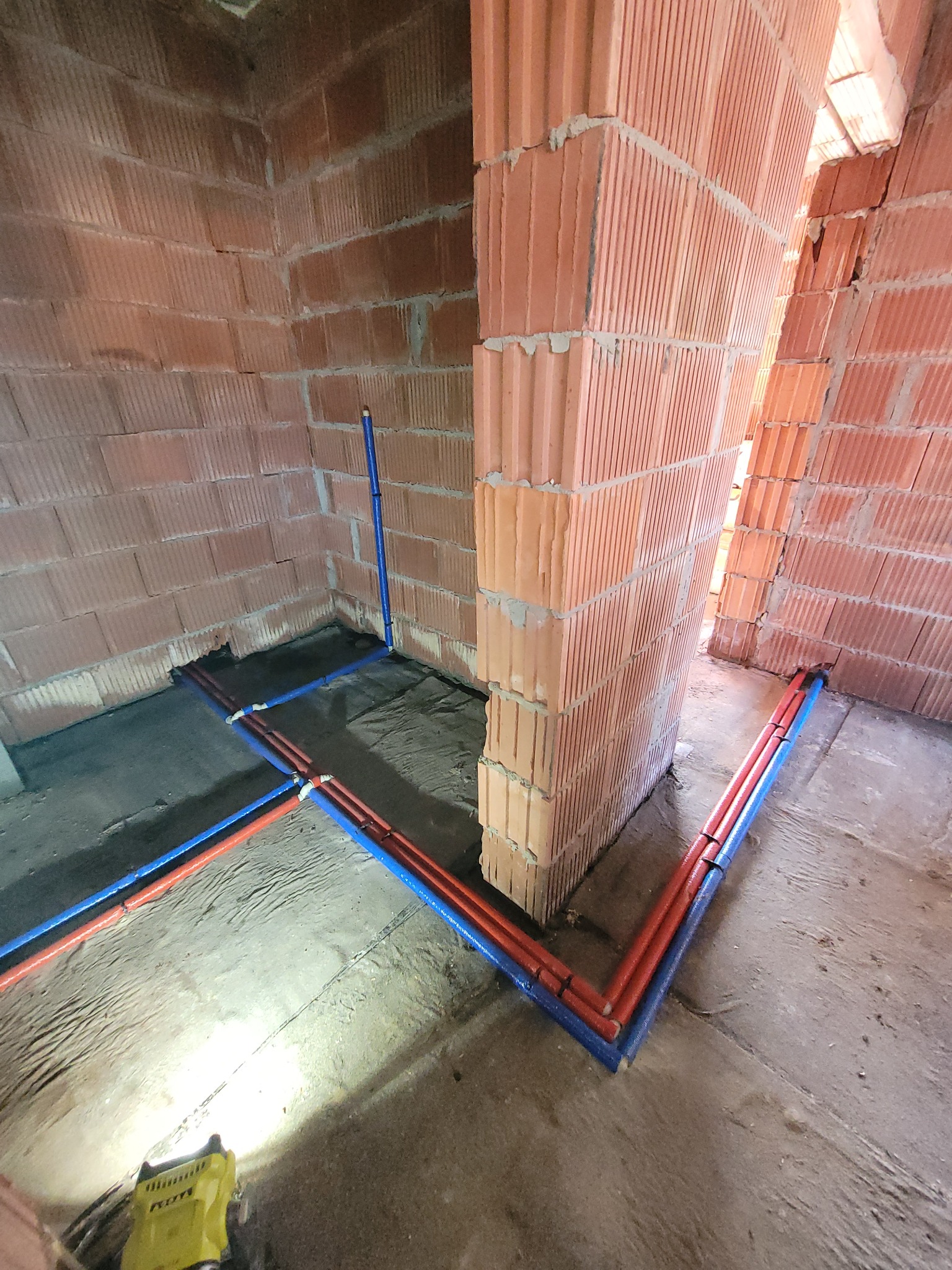 Instalacja wodna z rur PEX w kolorze niebieskim i czerwonym, poprowadzona po podłodze w surowym stanie, w pomieszczeniu z ceglanymi ścianami, oświetlona lampą roboczą