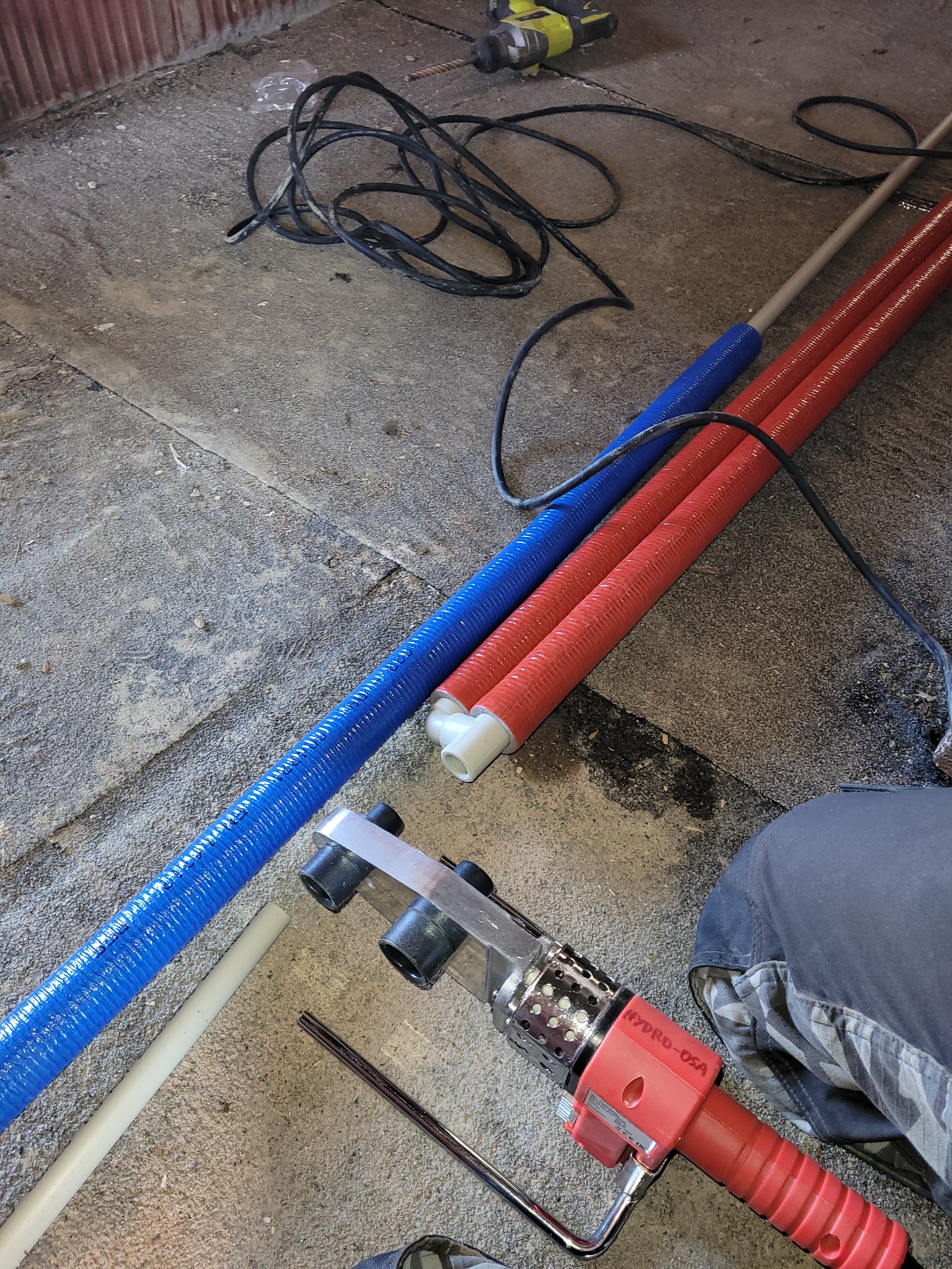 Przygotowanie do łączenia rur PEX: niebieska i czerwona rura izolowana termicznie leżą obok zgrzewarki HYDRD-OSA na betonowej posadzce, w tle wiertarka akumulatorowa.